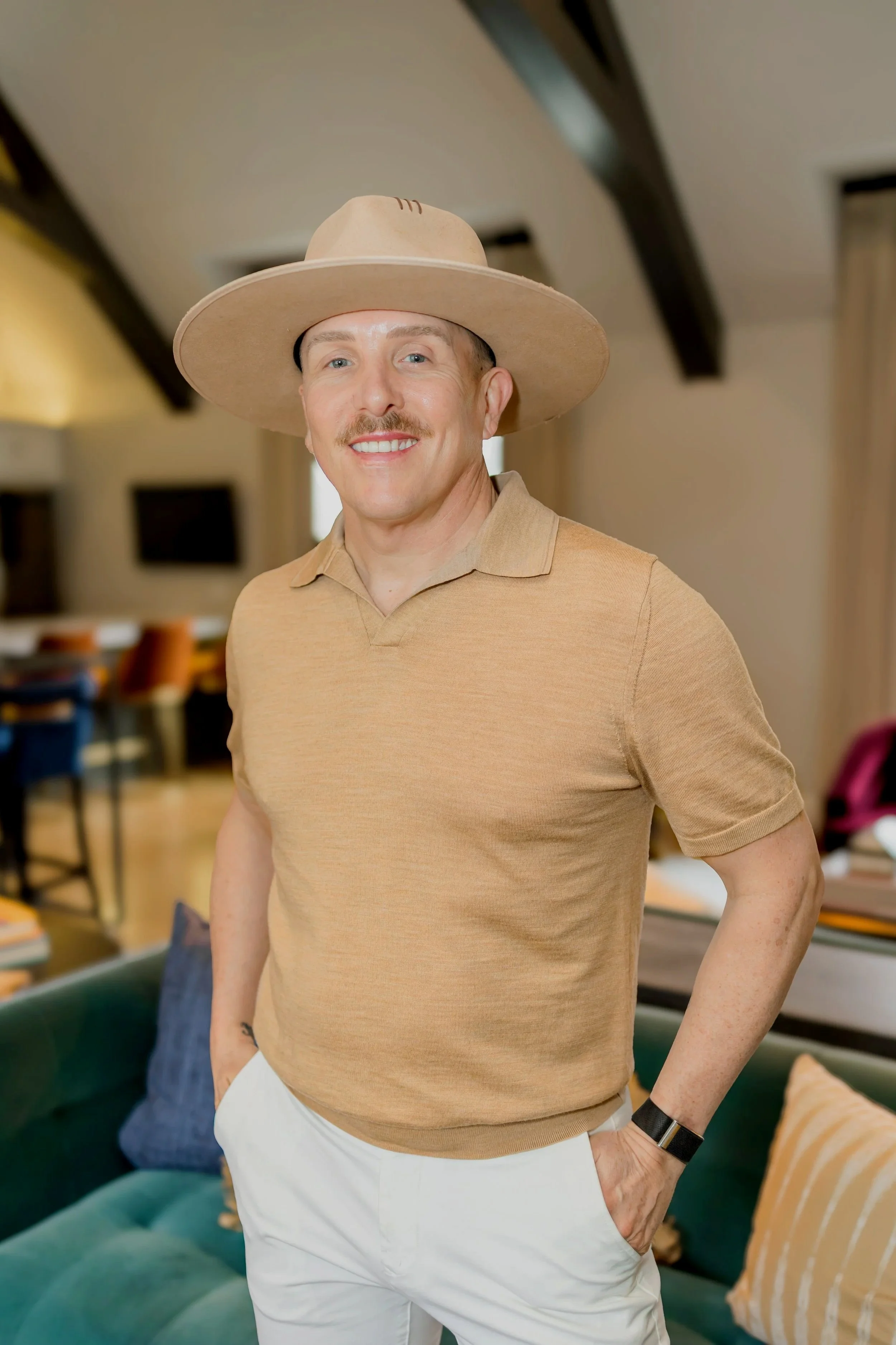A man with a tan complexion, wearing a beige wide-brimmed hat, a light brown polo shirt, and white pants, standing with hands in pockets in a cozy room with beige walls, wooden beams, colorful chairs, a couch, and windows.