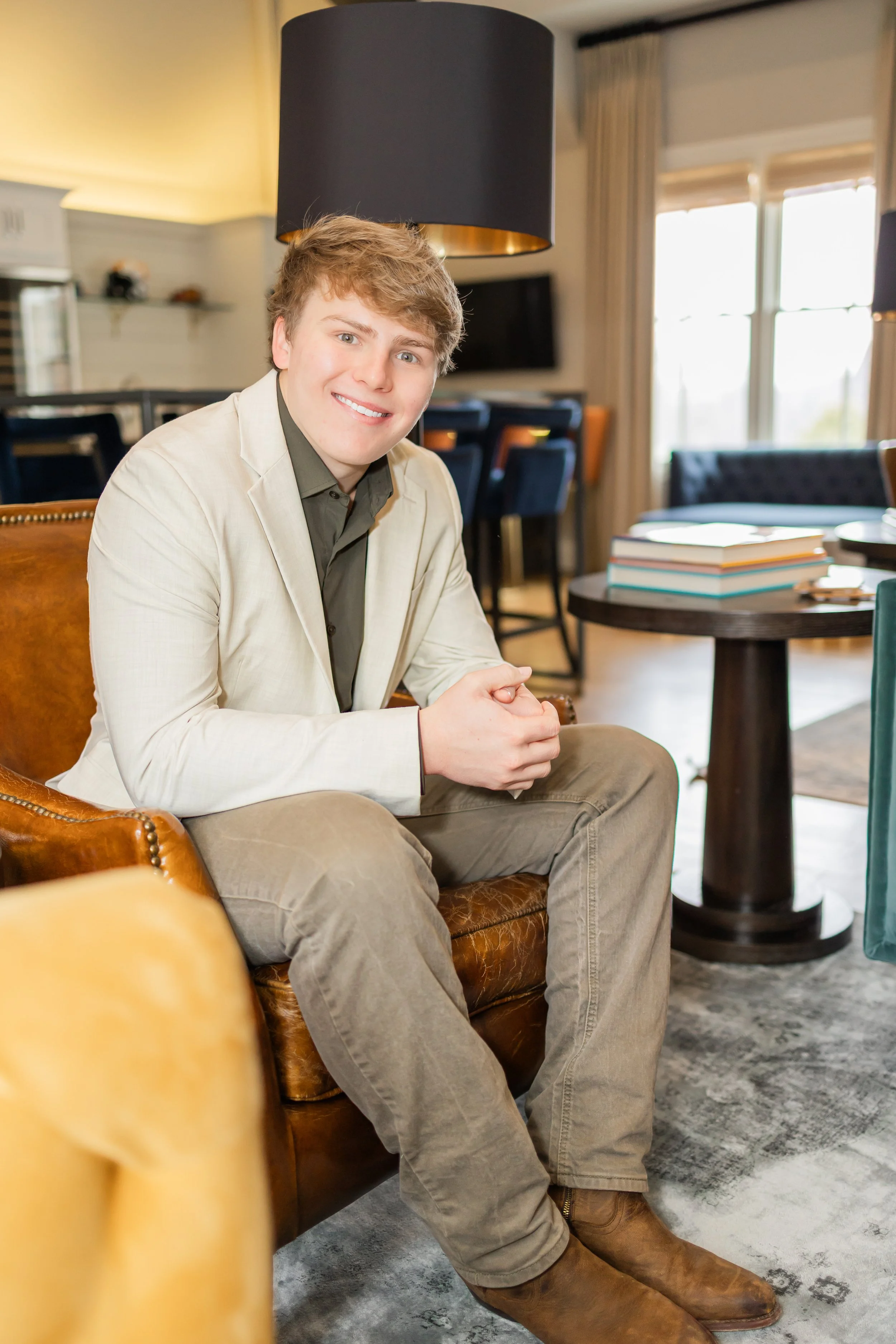 A young man with light brown hair, wearing a beige blazer, dark shirt, khaki pants, and brown boots, sitting on a leather armchair in a well-lit room with large windows, books on a dark round table, and a modern interior design.