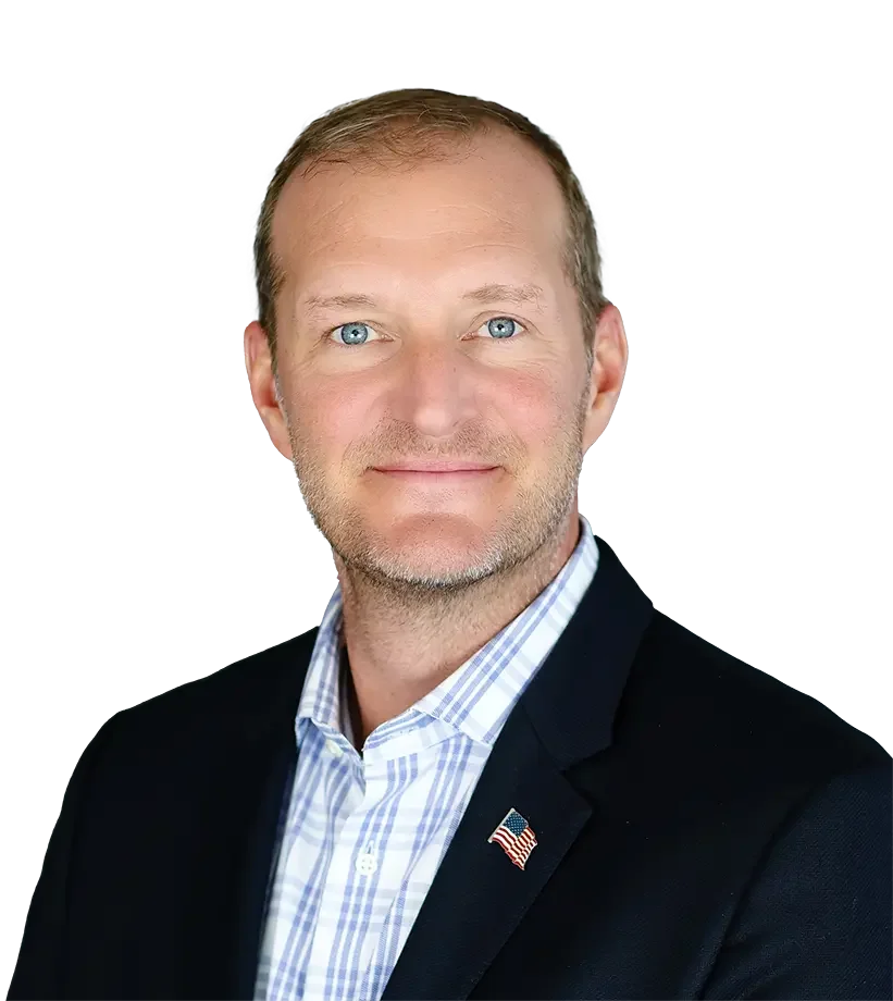 A portrait of a middle-aged man with blue eyes and light brown hair, wearing a dark blazer, a striped button-up shirt, and a small American flag lapel pin, against a plain background.