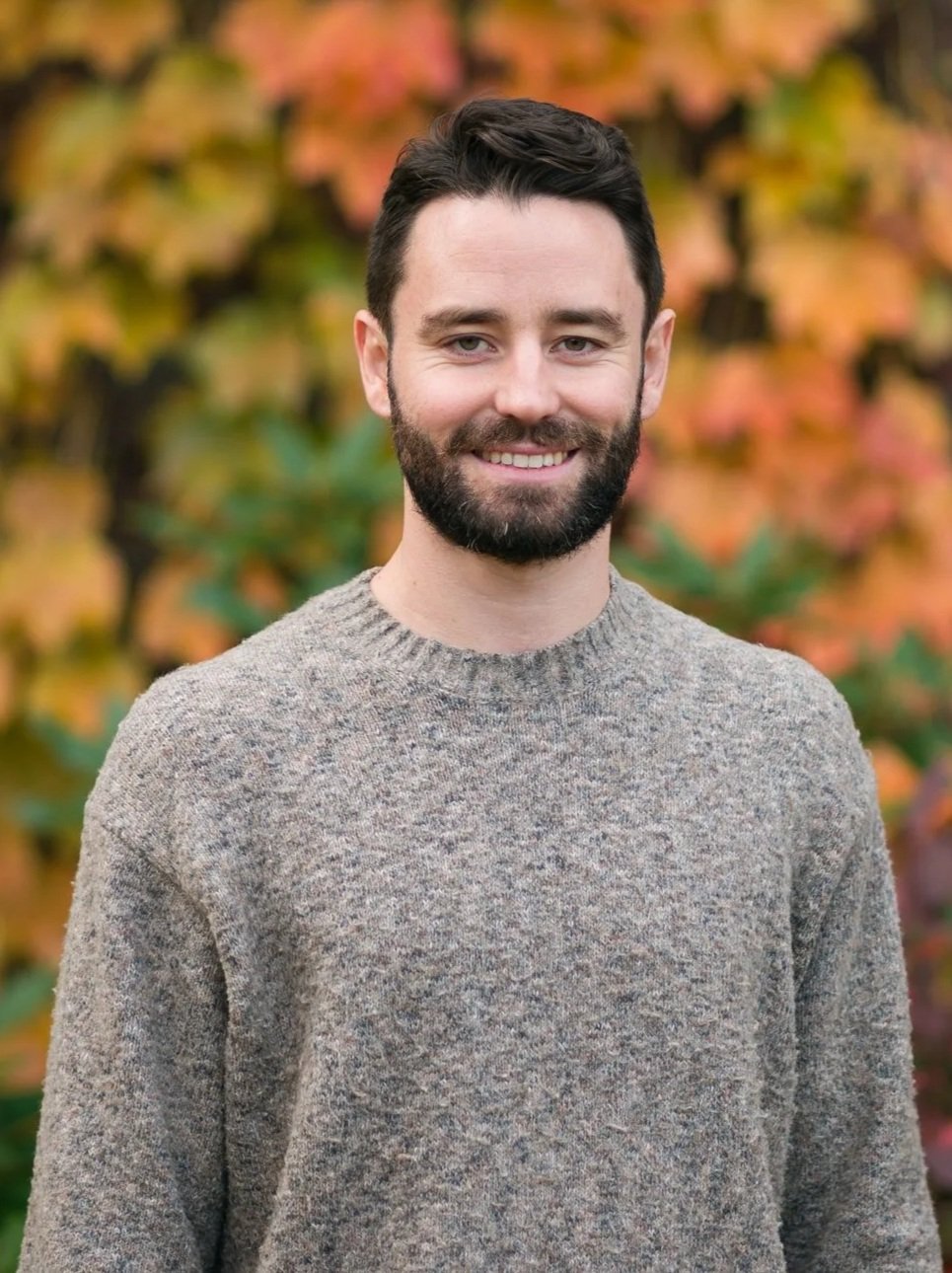 A man with dark hair and a beard standing outdoors in front of a brick building and colorful autumn foliage.