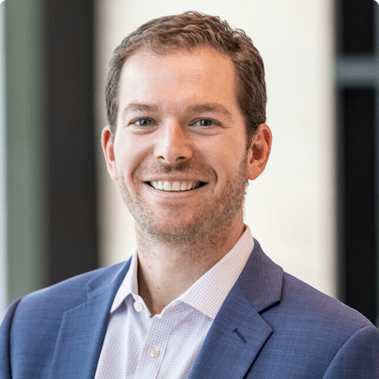 A smiling man in a blue business suit and white shirt, standing indoors with a blurred background.