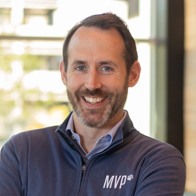 Man with short brown hair, beard, and blue eyes smiling, wearing a blue sweater with a logo on the chest, standing indoors with large windows behind him.