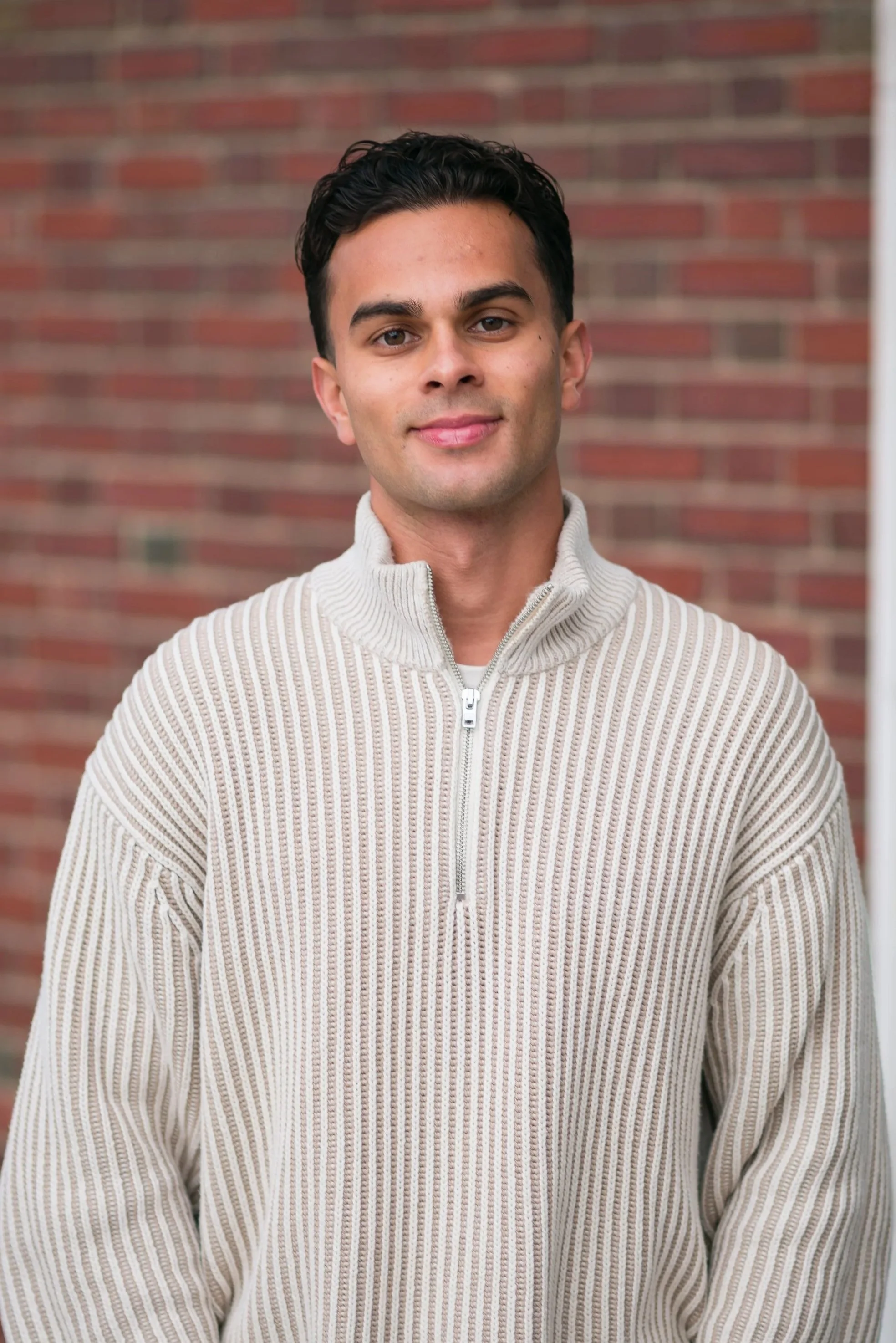 A young man with dark hair wearing a beige zip-up sweater and black pants, standing outdoors on a sidewalk with trees and houses in the background.