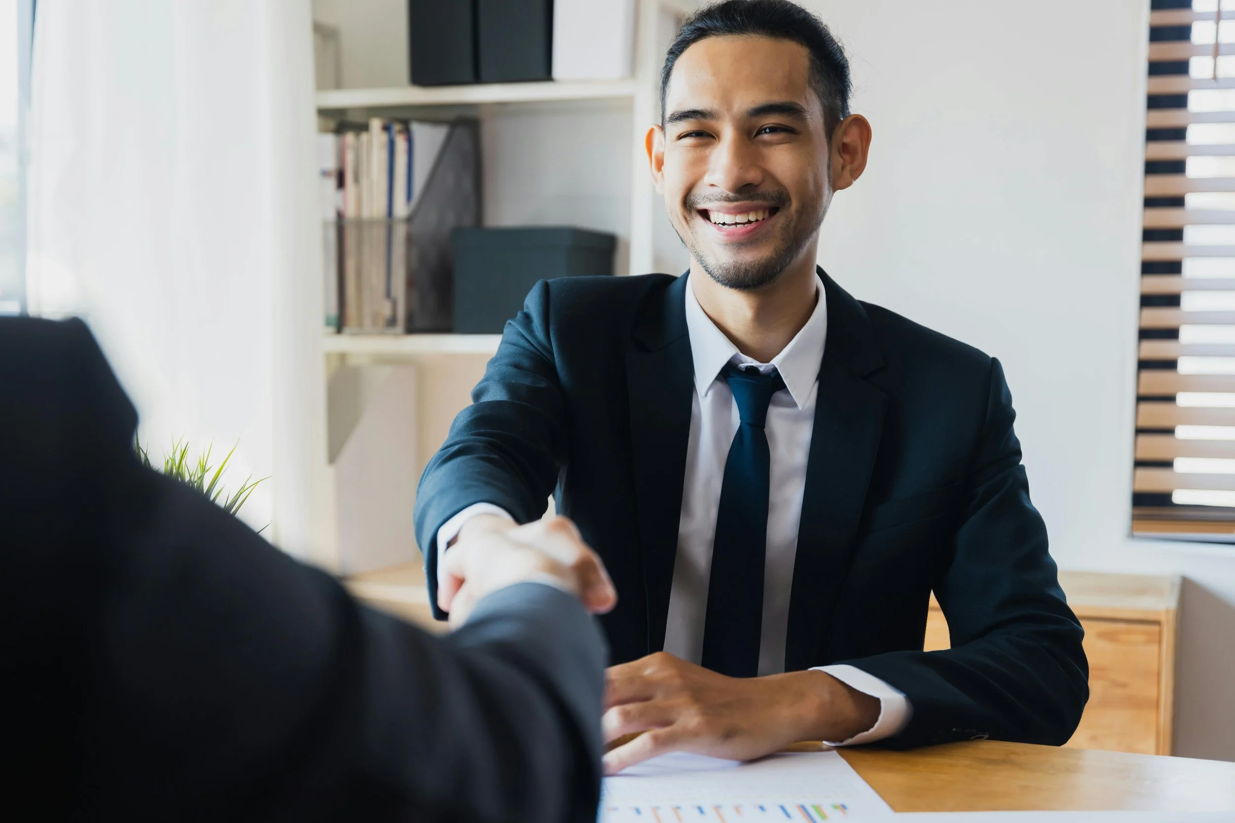 A man in a business suit shaking hands with another person across a wooden table in an office setting.