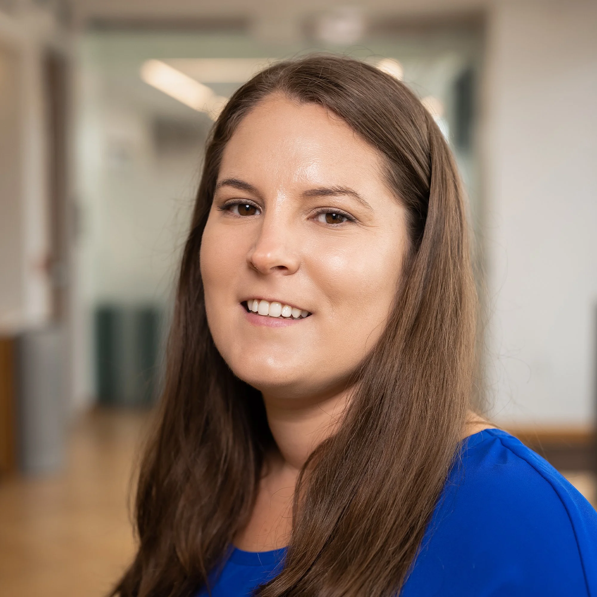 Portrait of a woman with long brown hair wearing a blue top, smiling in an indoor setting.