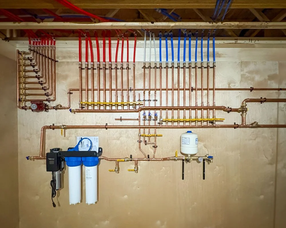 A plumbing system with copper pipes, red and blue PEX tubing, a water filter, and control valves installed on a unfinished wooden wall.