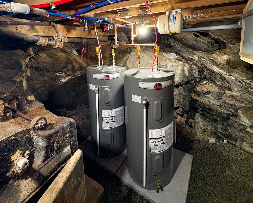 Two Rheem water heaters in a basement with exposed rock walls and plumbing pipes overhead.