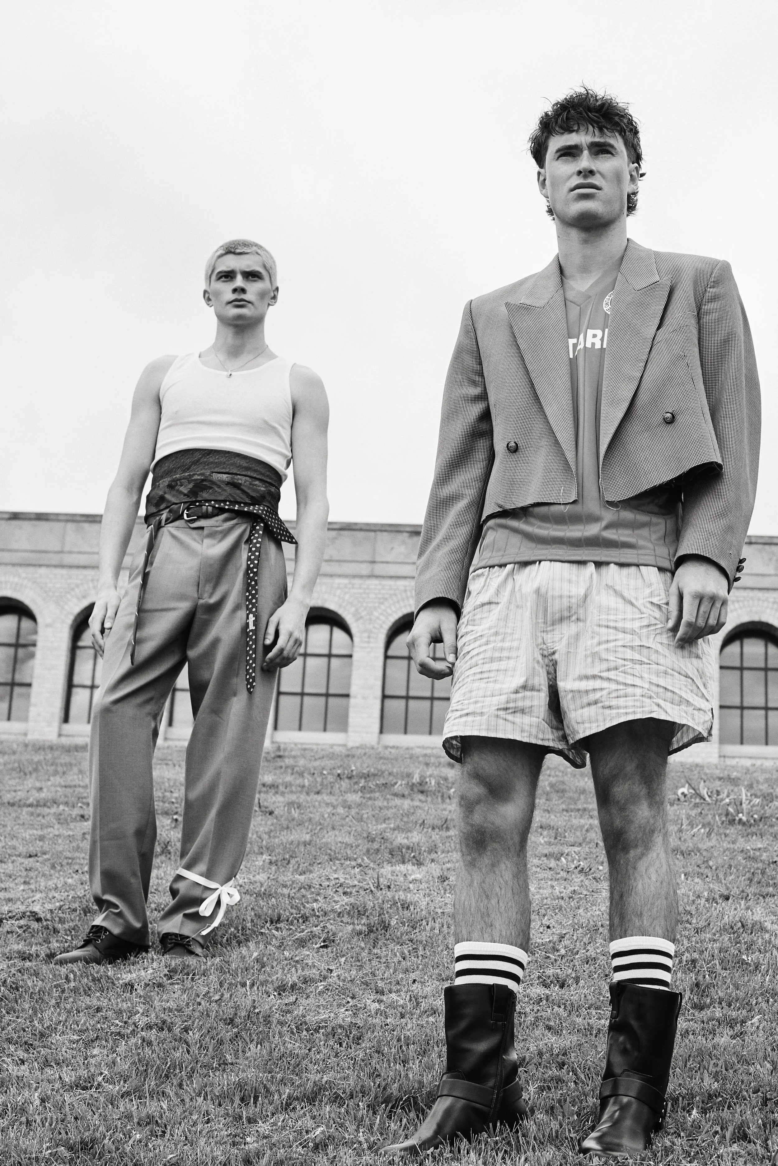 Two young men standing on grass in front of a building with arched windows, dressed in casual and eclectic fashion, black and white photo.