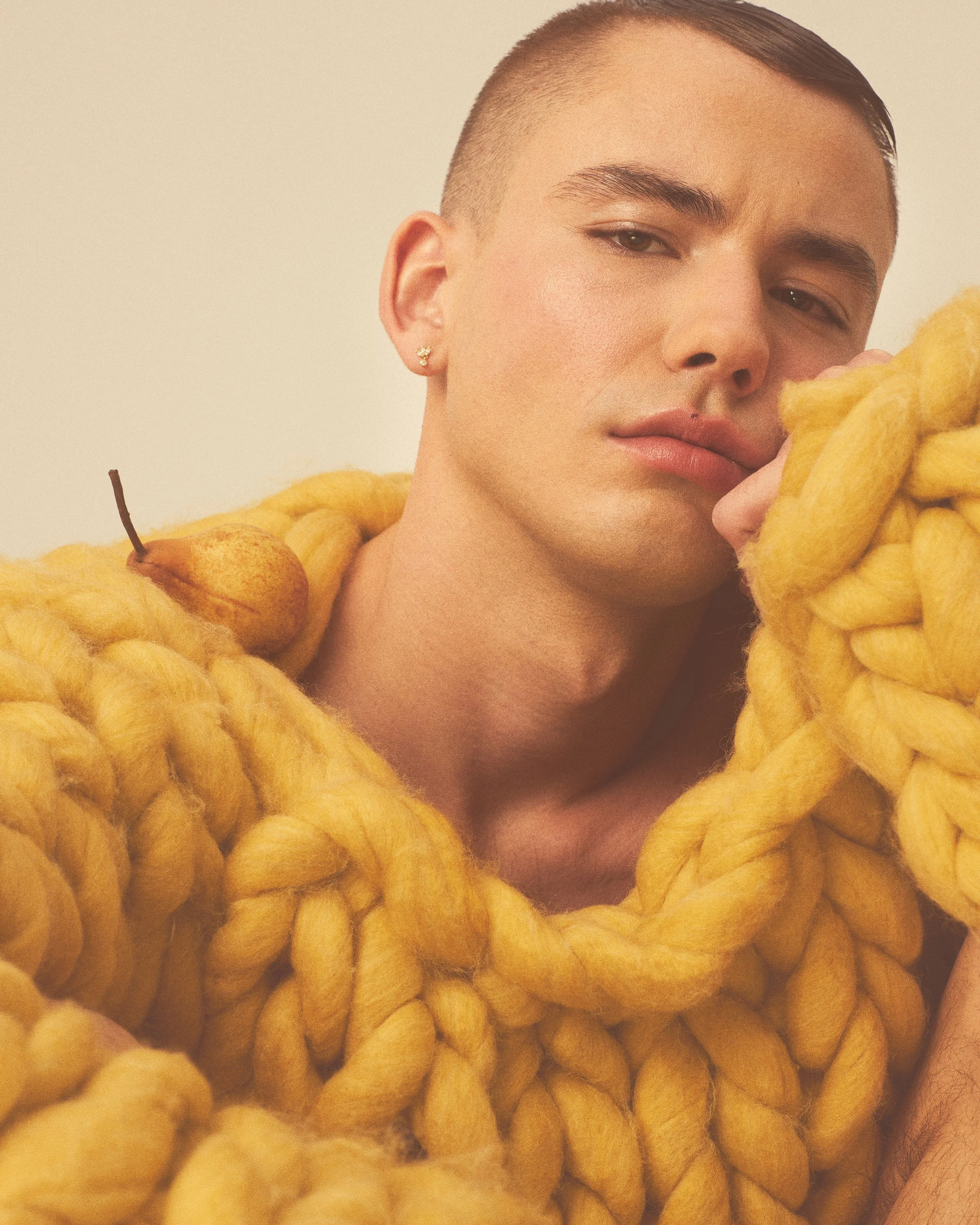A young person with short hair, wearing a large yellow knitted garment with a pear perched on the shoulder, against a plain background.