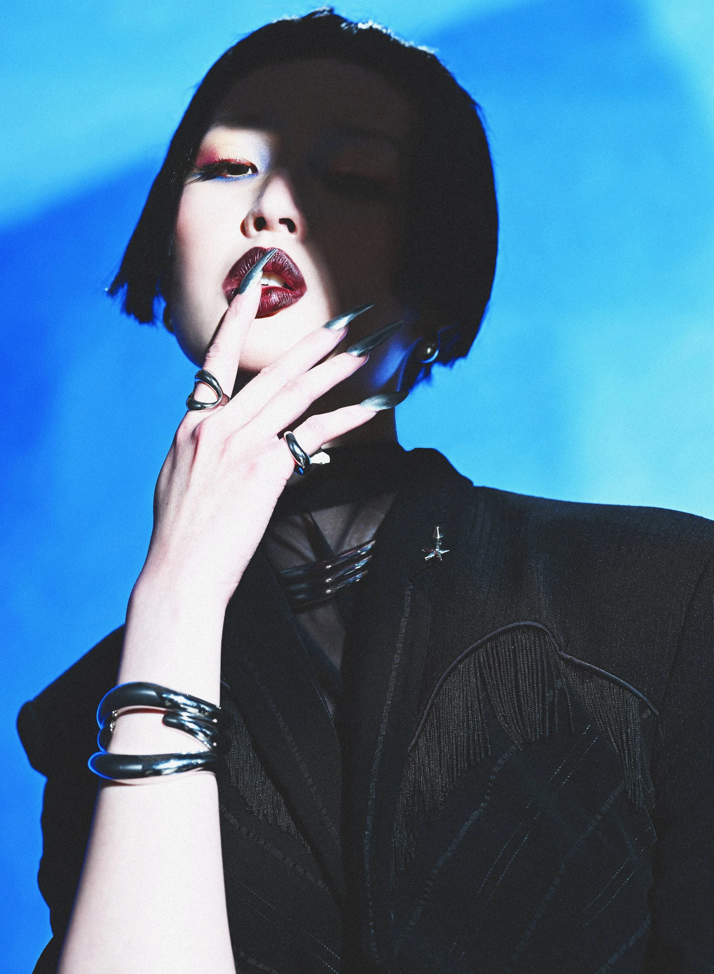 A woman with short black hair and makeup posing against a blue background, wearing dark clothing and jewelry, with her hand near her face.