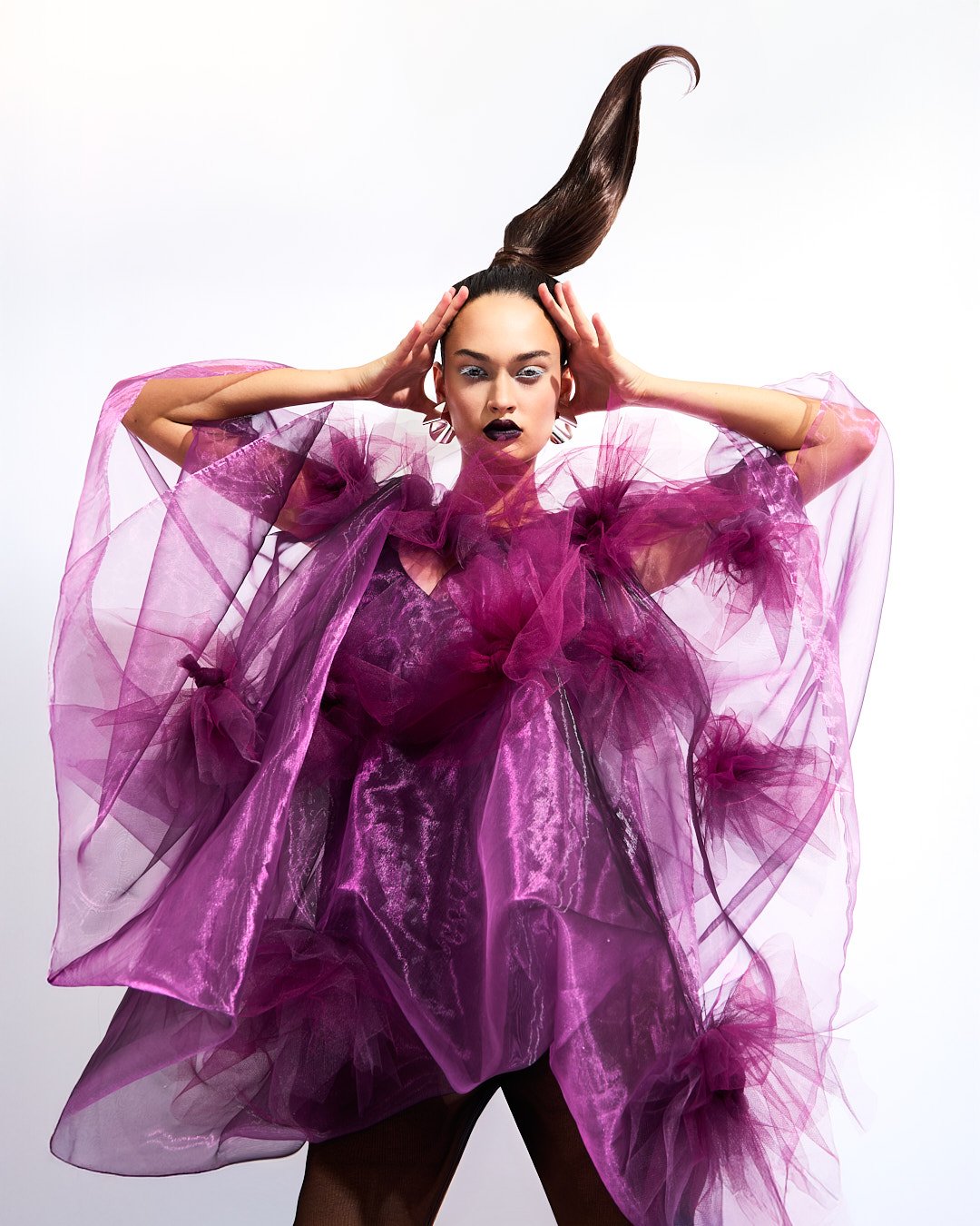 A fashion model wearing an elaborate purple dress with tulle and ruffles, posed with hands on her head and hair styled upward.