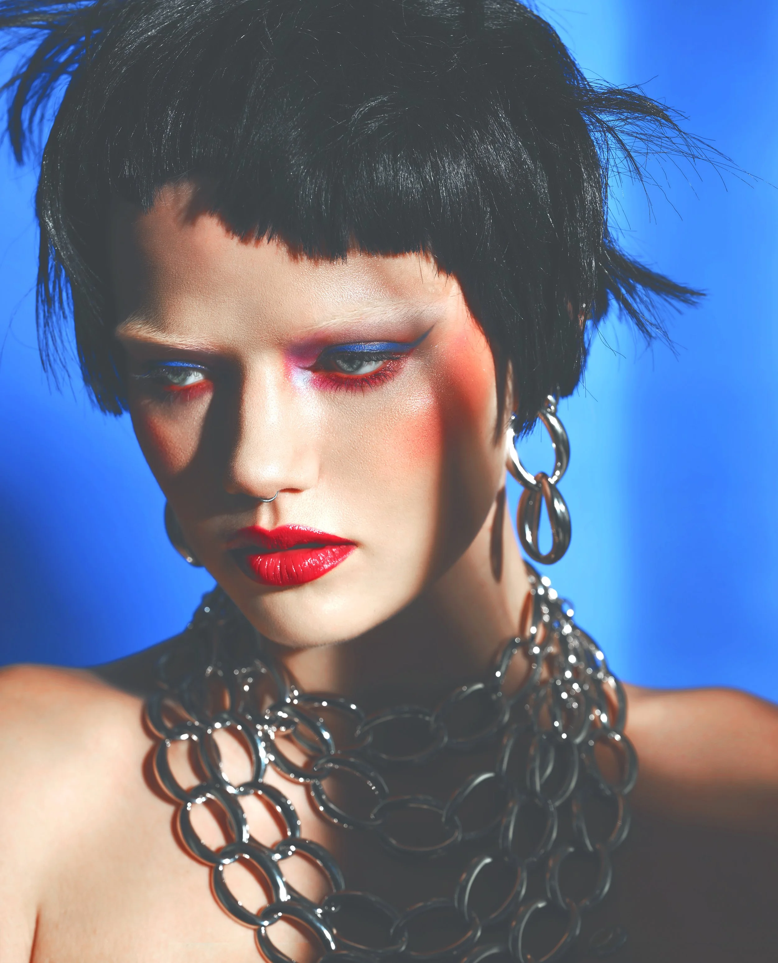 A woman with short black hair, bold makeup with pink, blue, and red eyeshadow, and red lipstick. She is wearing large silver hoop earrings and layered silver chain necklaces against a blue background.