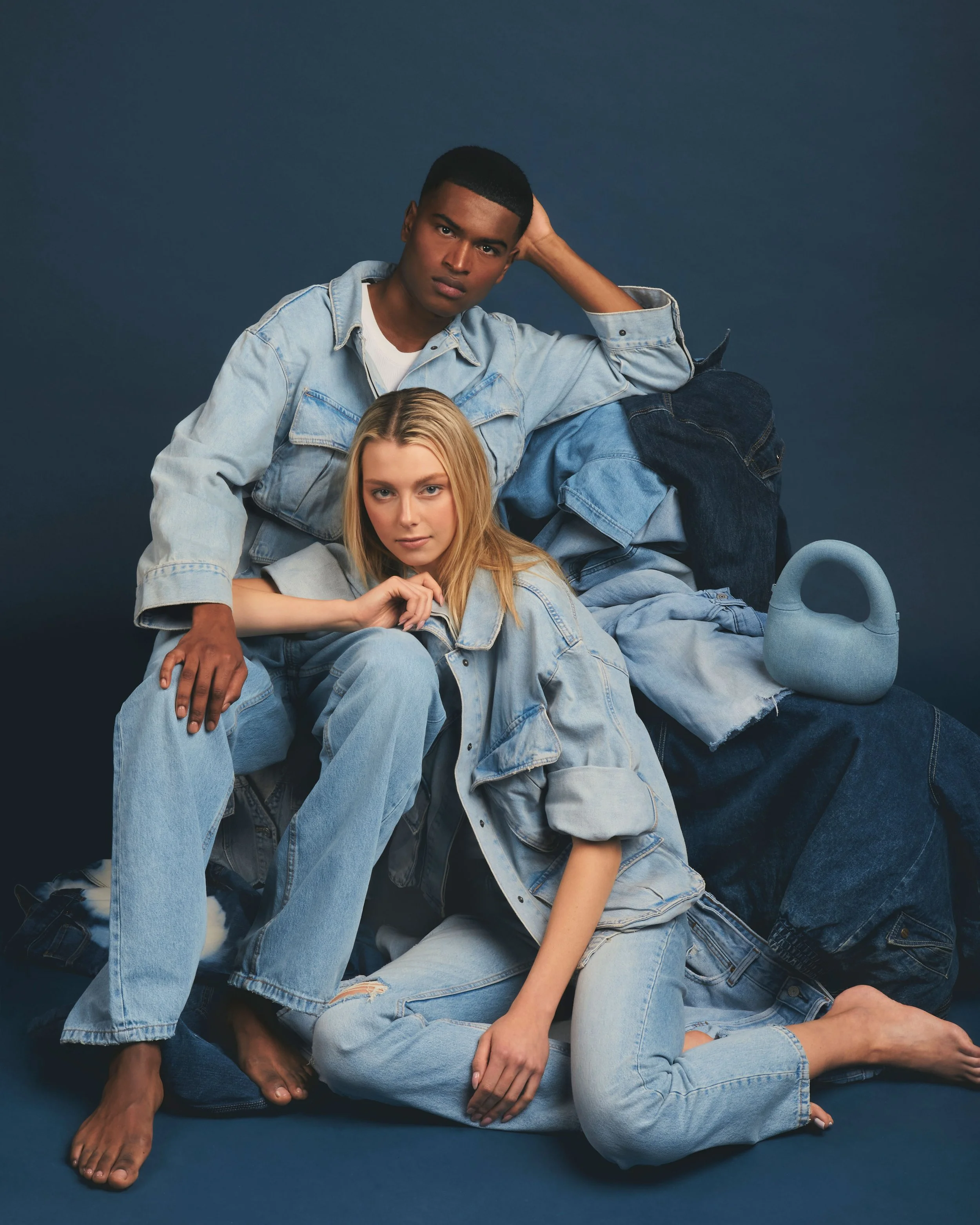 Two models, one male and one female, posing in denim outfits against a dark background, with a denim handbag nearby.