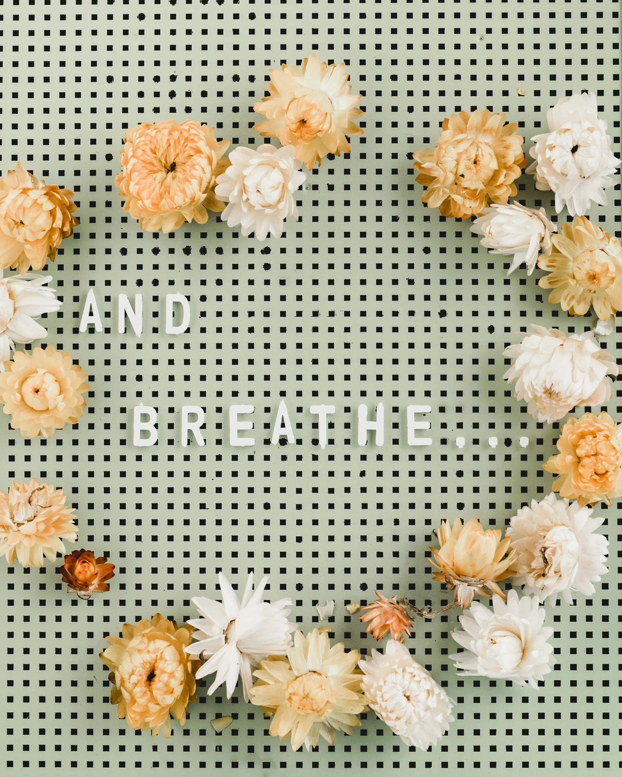 A decorative board with the words 'AND BREATHE' spelled out in white letters, surrounded by beige, white, and peach flowers on a black and white checkered background.
