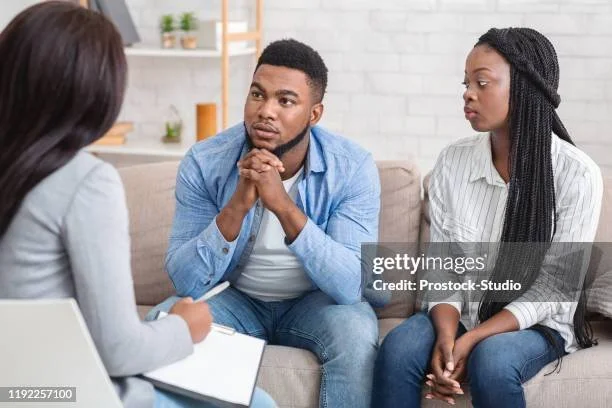 A man and woman sitting on a couch during a counseling session with a therapist, who is taking notes.