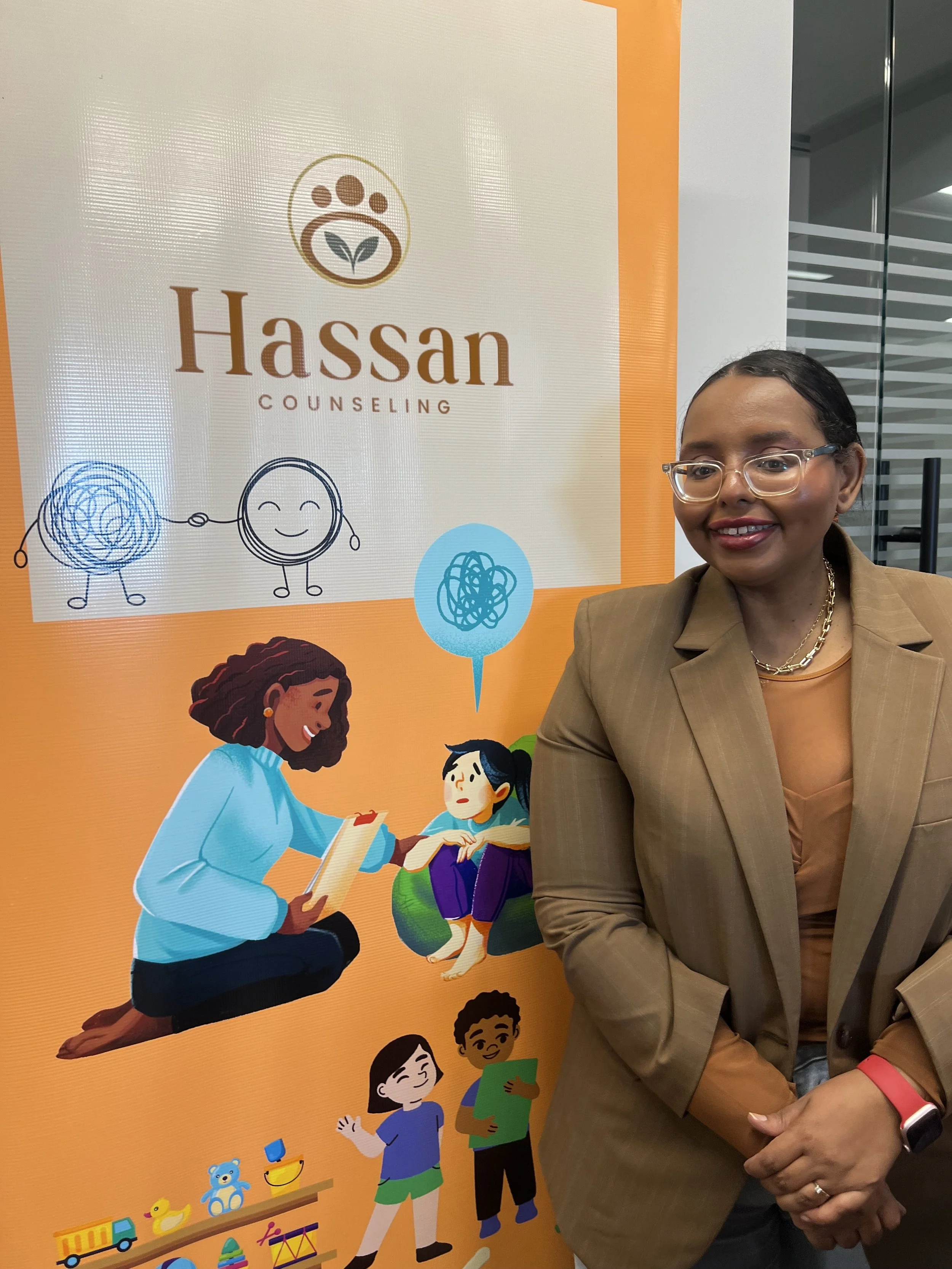 A woman standing in front of a sign for Hassan Counseling, which has cartoon illustrations of children and toys.