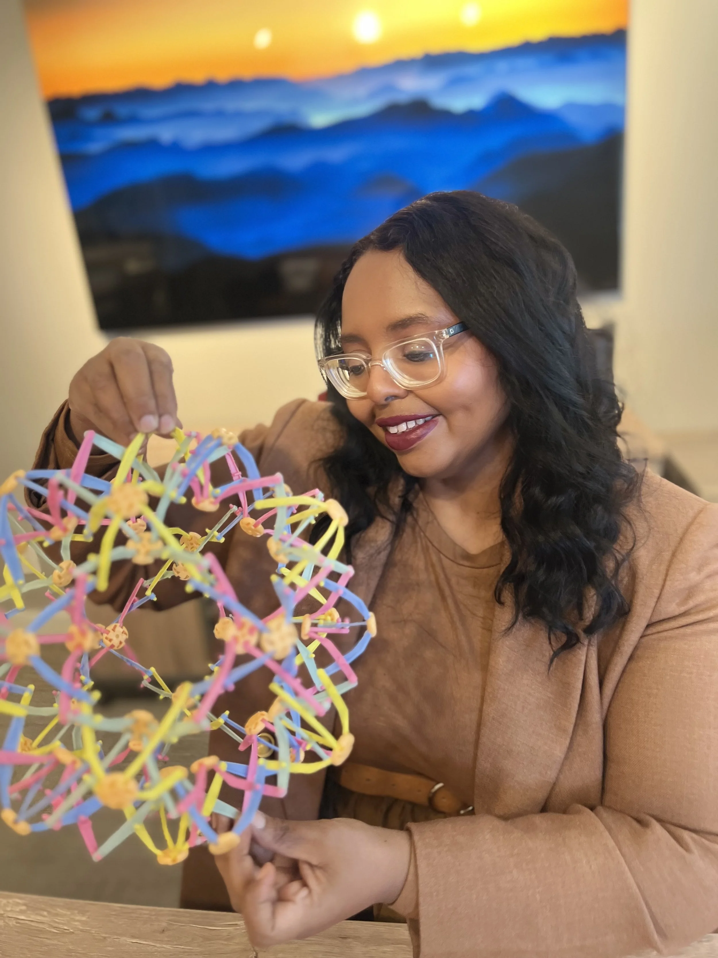 A woman with black wavy hair and glasses smiling as she holds a colorful molecular model with pink, yellow, blue, and green sticks and small orange connector pieces, with a sunset landscape painting in the background.