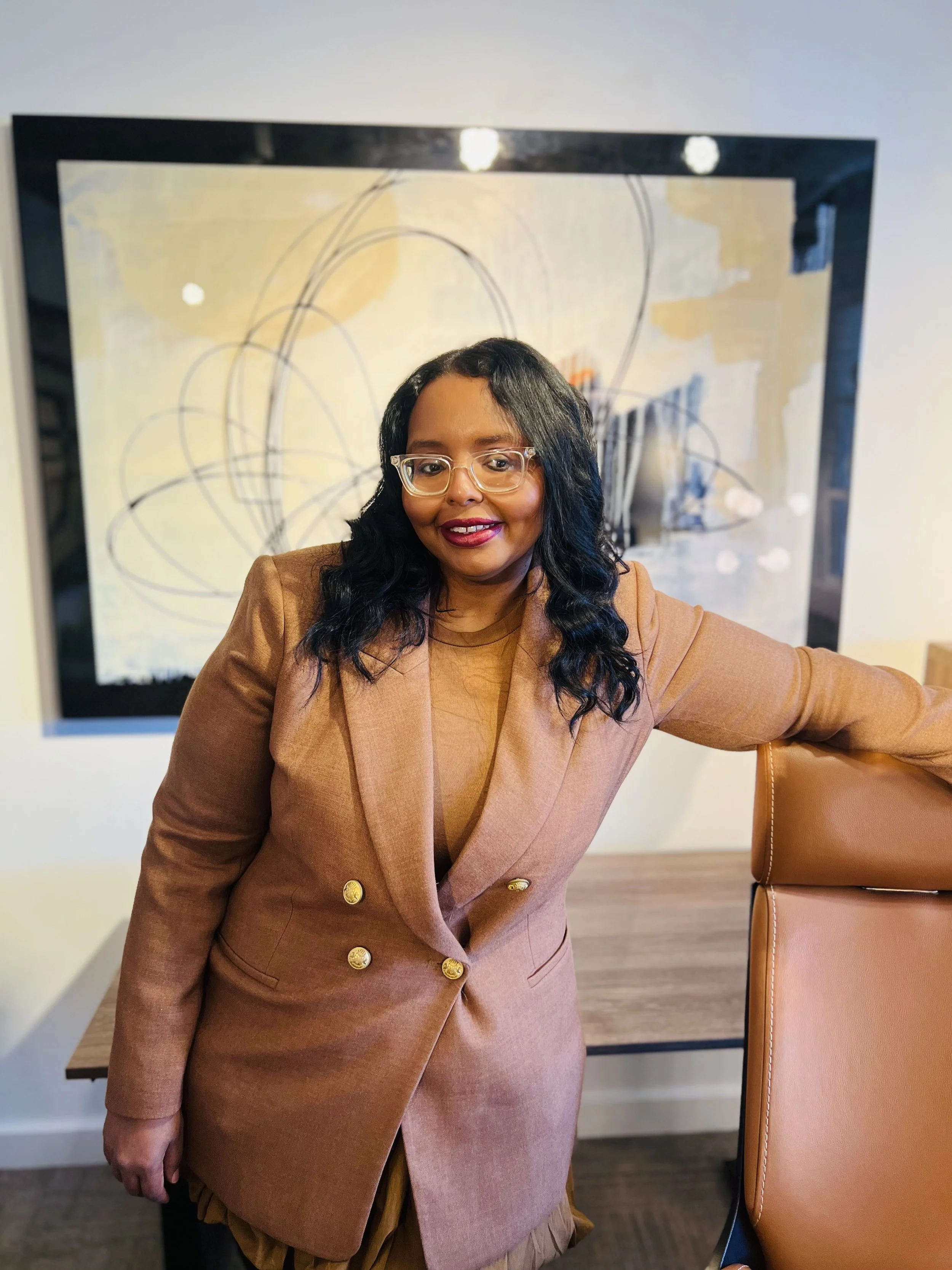 A woman with black, wavy hair wearing glasses and a brown blazer standing in an office or conference room, with a large painting featuring abstract loops and lines behind her.