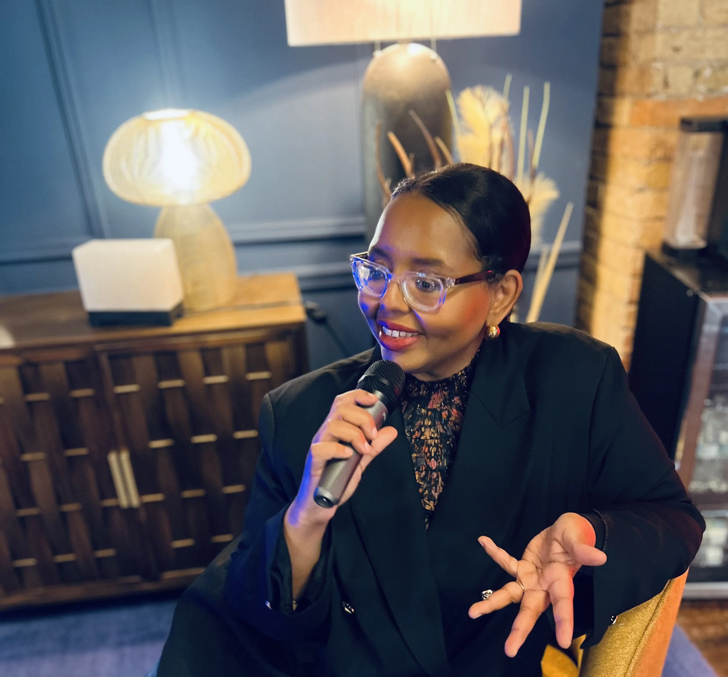 A woman with short black hair, glasses, and earrings speaking into a microphone while seated in a warmly lit room with blue walls and wooden furniture.