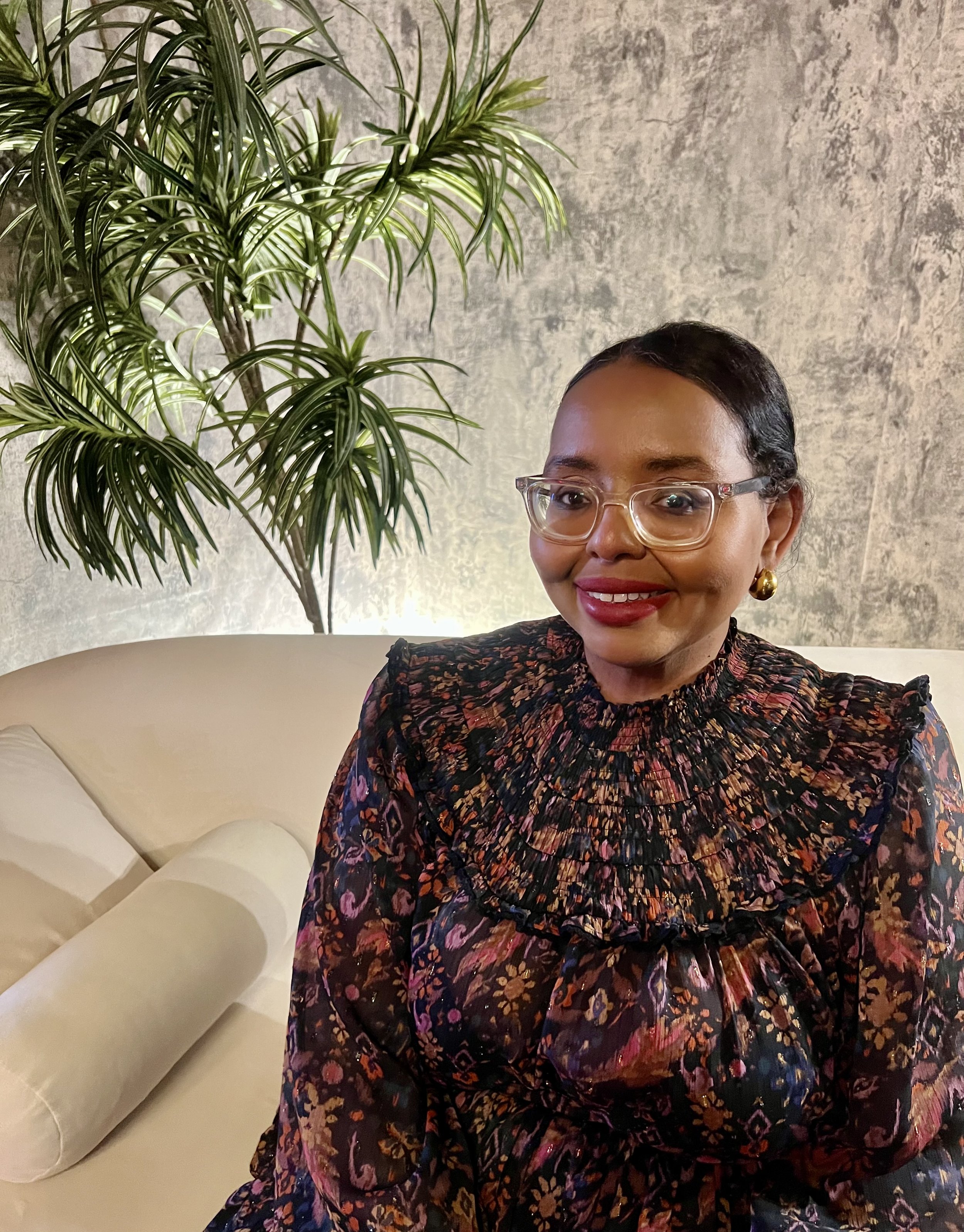 A woman with short dark hair, clear glasses, red lipstick, gold earrings, smiling, sitting on a beige couch, wearing a dark floral blouse, in front of a textured gray wall and a large green plant.