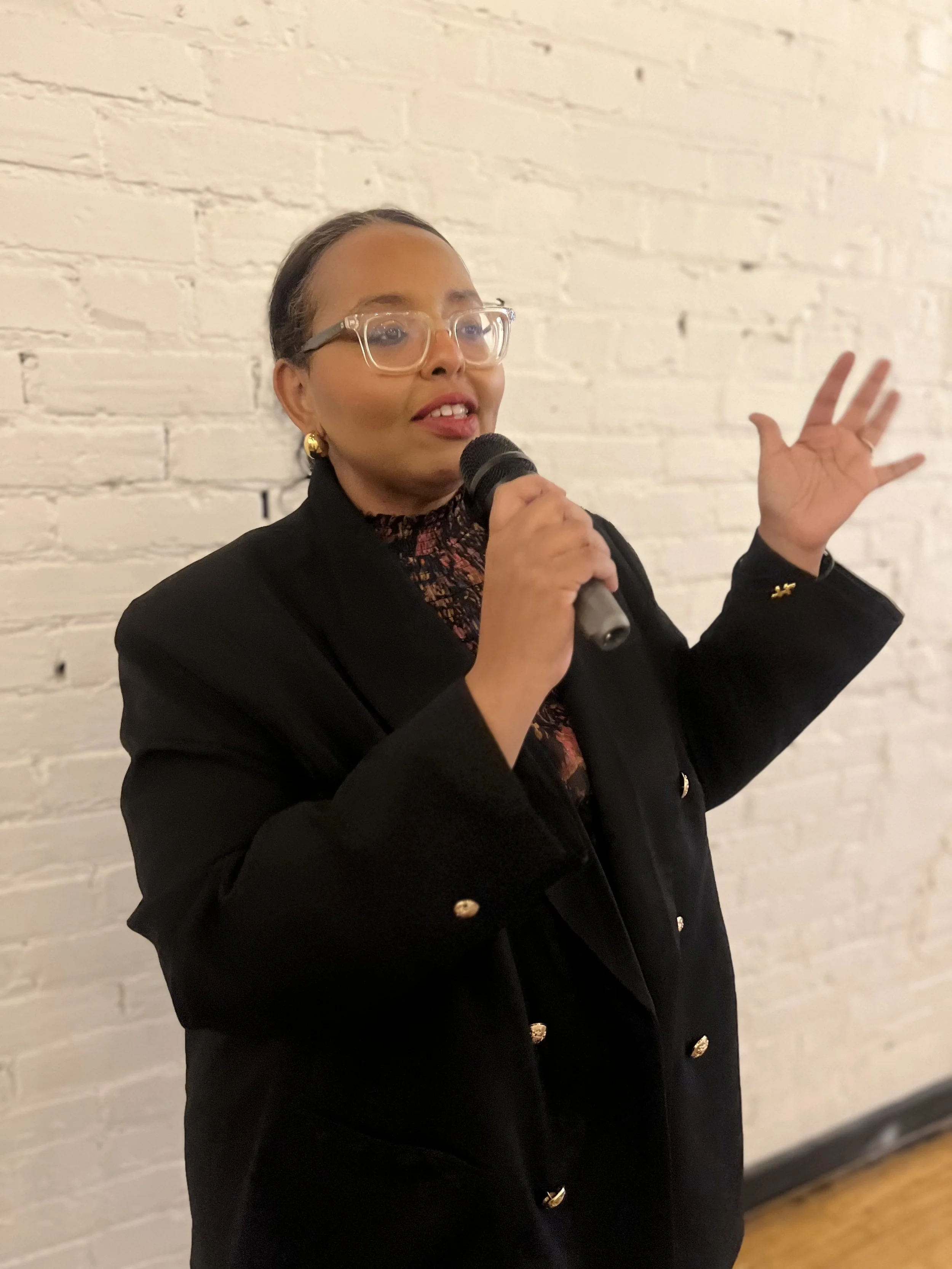 A woman with glasses and earrings speaks into a microphone against a white brick wall.