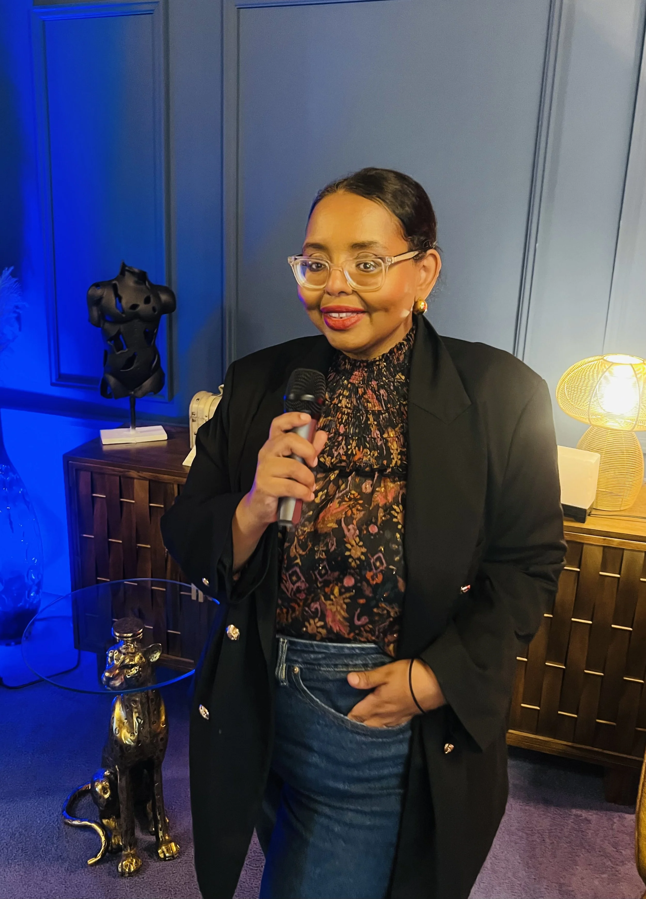 A woman with glasses and earrings holding a microphone, wearing a black blazer and floral blouse, standing in a room with blue walls and warm lighting, a decorative statue of a black cat on a side table.