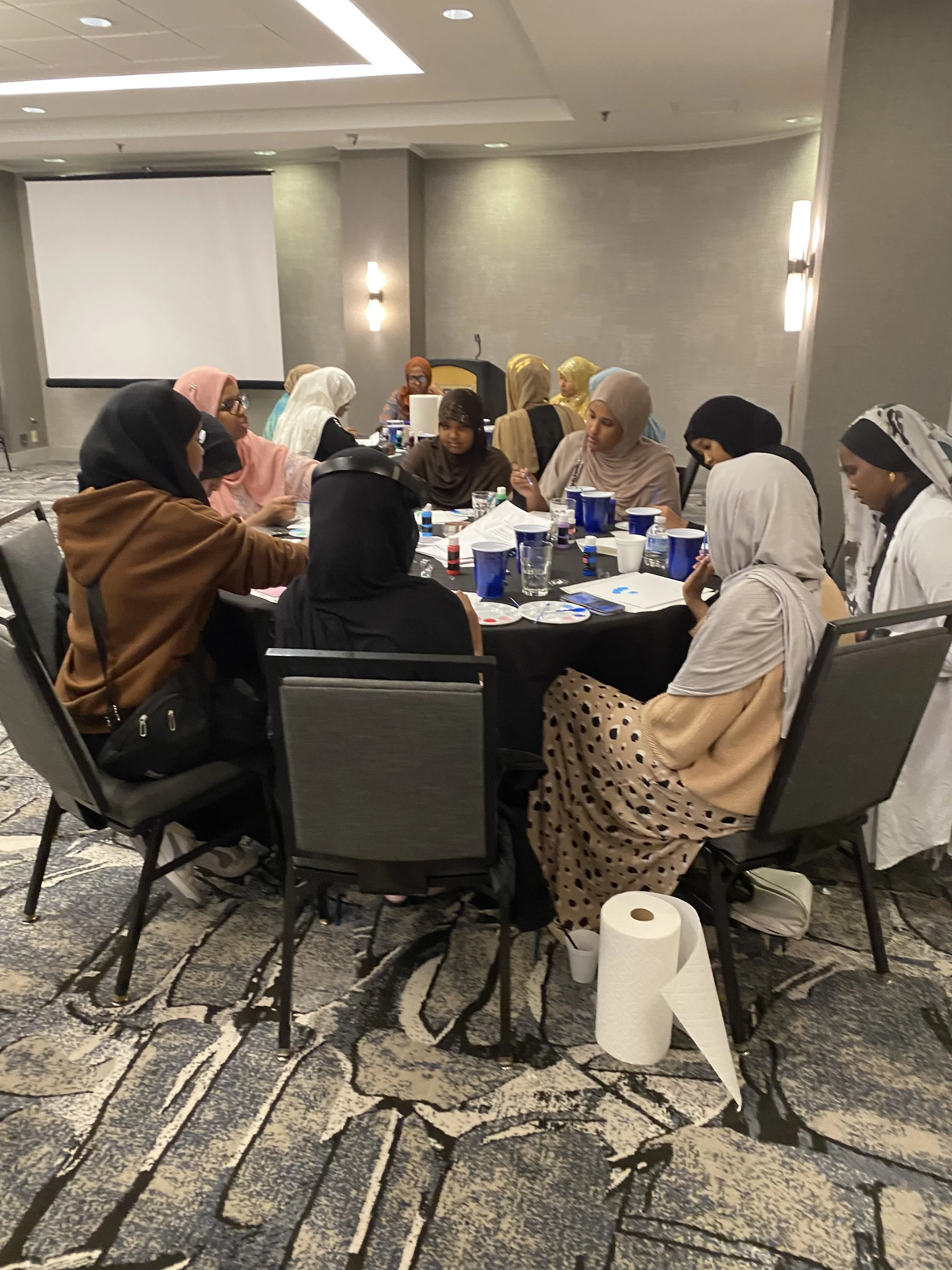 A group of women sitting around a table, engaged in arts and crafts activities in a conference room with a large screen and patterned carpet.
