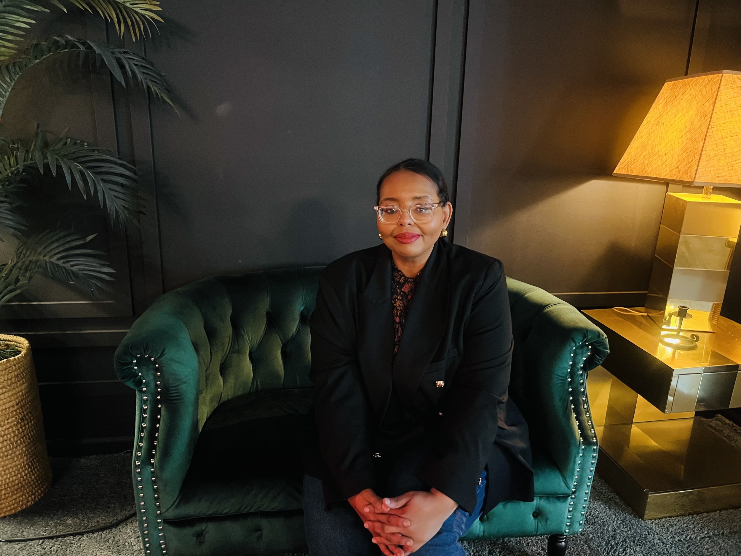 A woman sitting on a green velvet sofa in a dimly lit room with a black background, potted plant on the left, and a table with a lamp on the right.