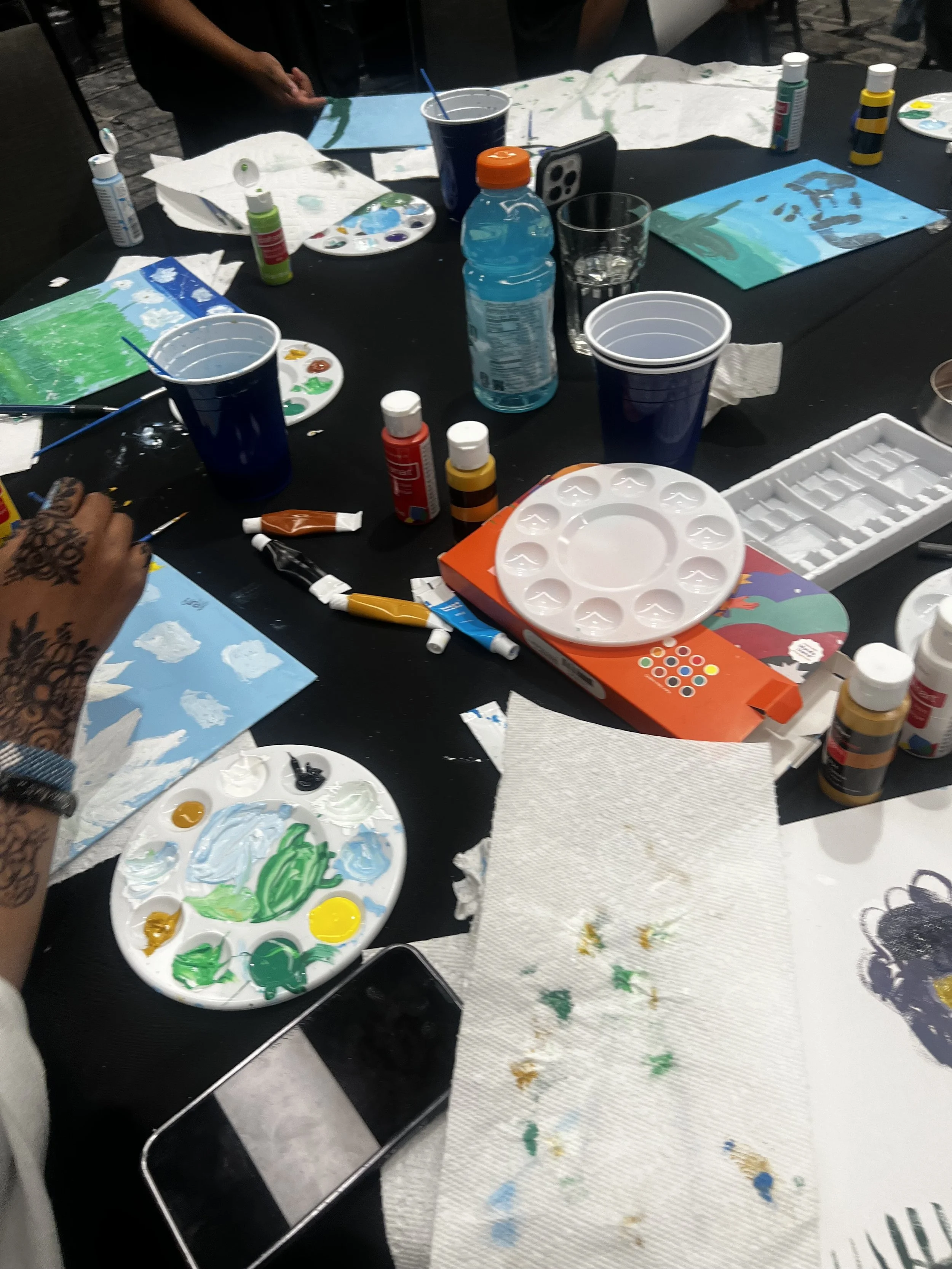 Table with art supplies, including paint palettes, paint bottles, brushes, paper, and cups, in the midst of a painting activity.