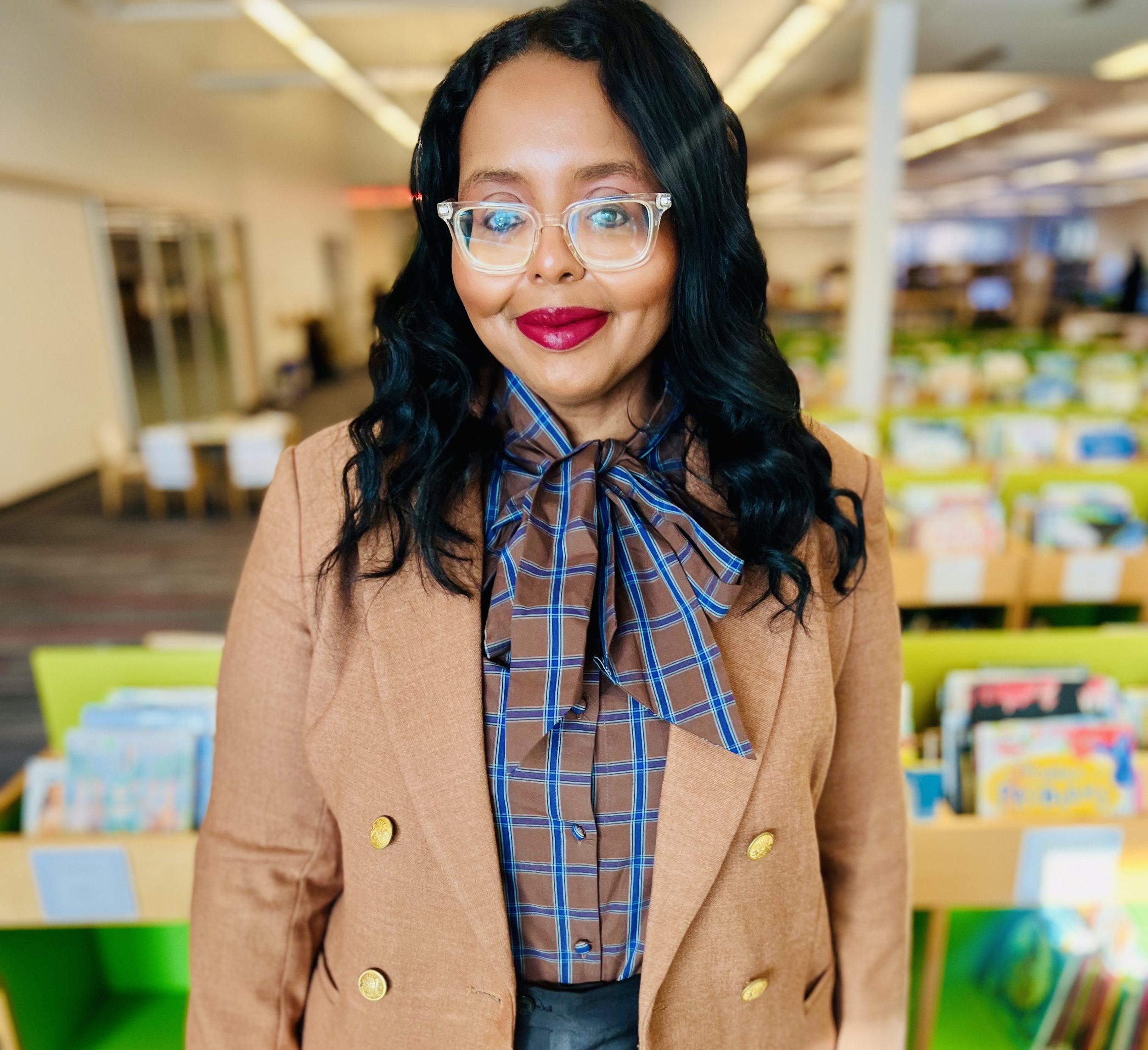A woman with black hair, wearing clear glasses, red lipstick, a brown blazer with gold buttons, and a plaid bow tie, standing in a colorful classroom or library with bookshelves.