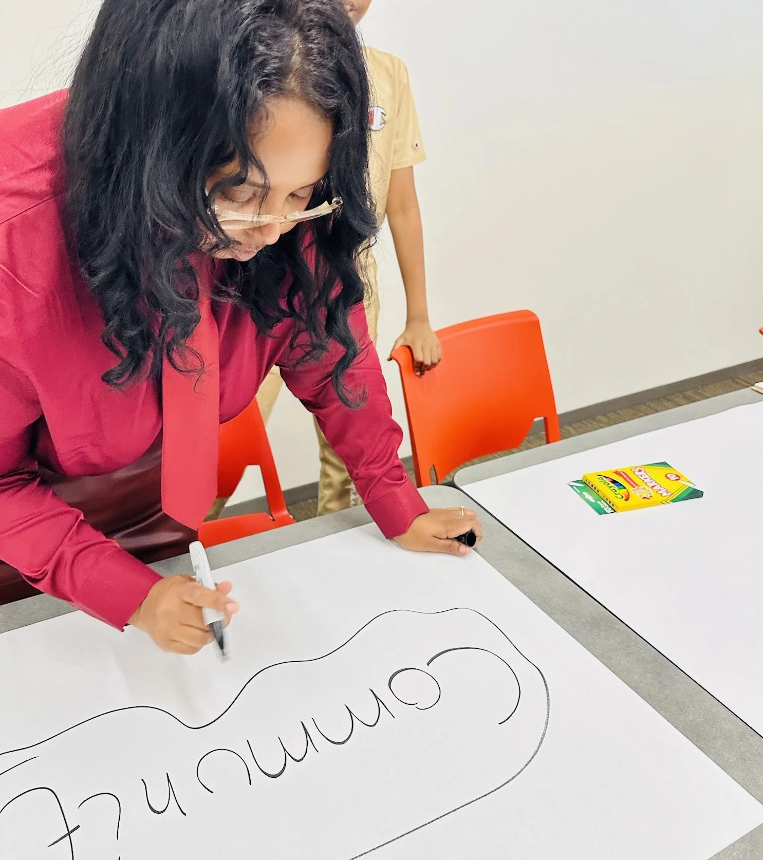 A woman in a pink shirt and glasses is writing on a large white paper with a black marker. There is another person partially visible in the background, standing behind a red chair. Crayons are on the table.