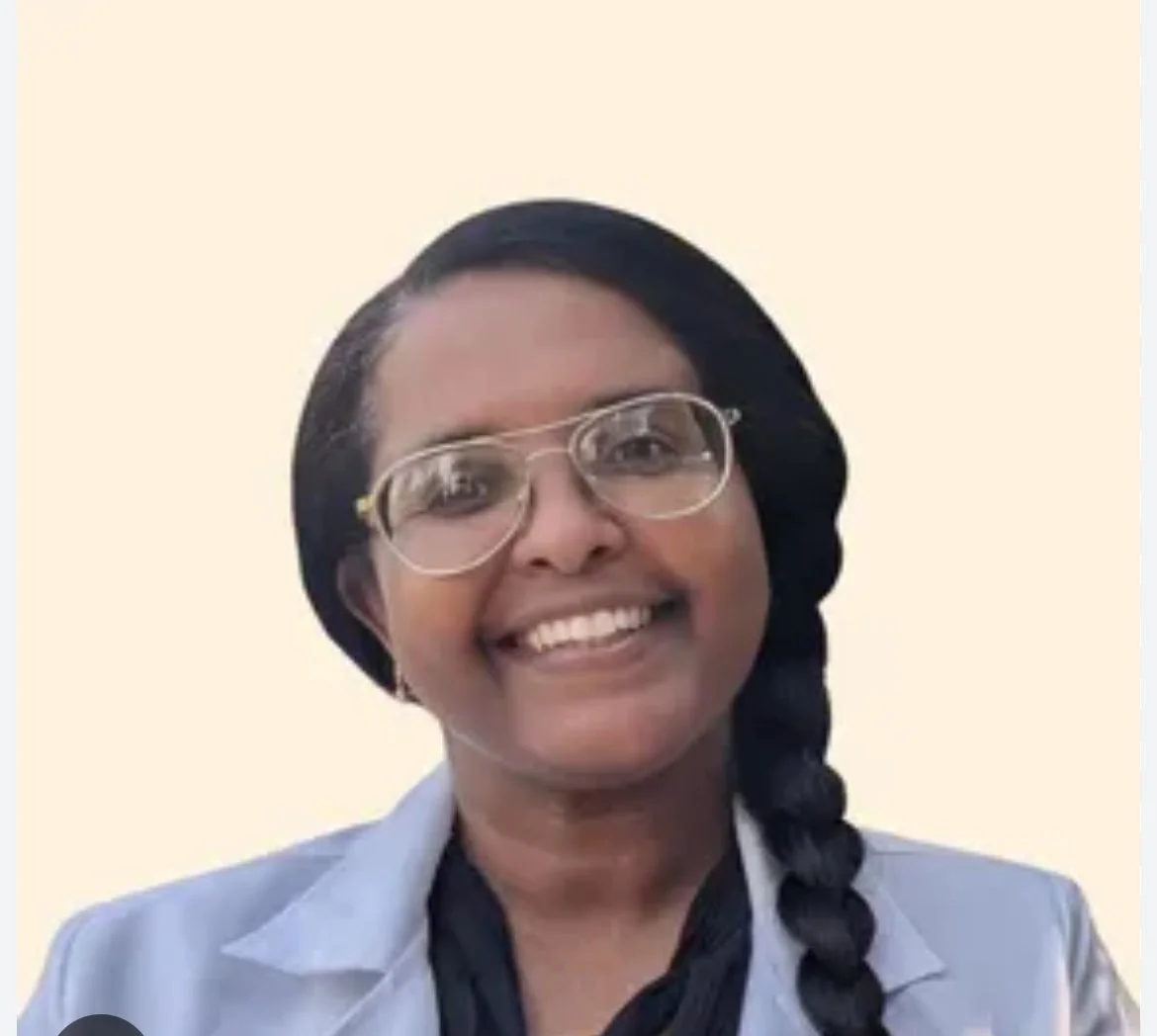 Smiling woman with glasses, black hair in braid, wearing a gray blazer and black shirt against a beige background.