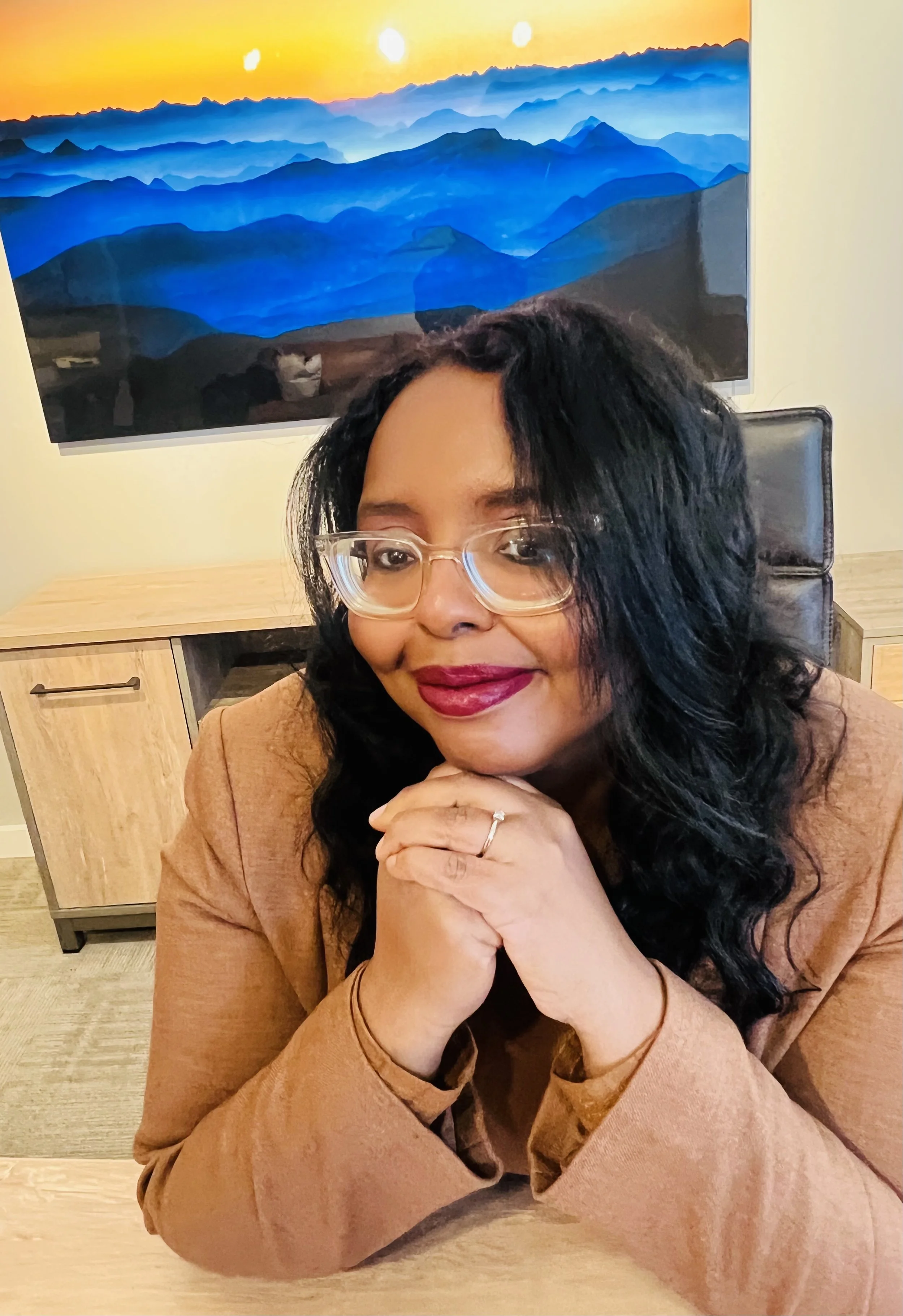 A woman with glasses and wavy black hair, wearing a tan blazer, sitting at a desk with her hands clasped under her chin, smiling at the camera. Behind her is a large framed picture of a mountain landscape at sunset.