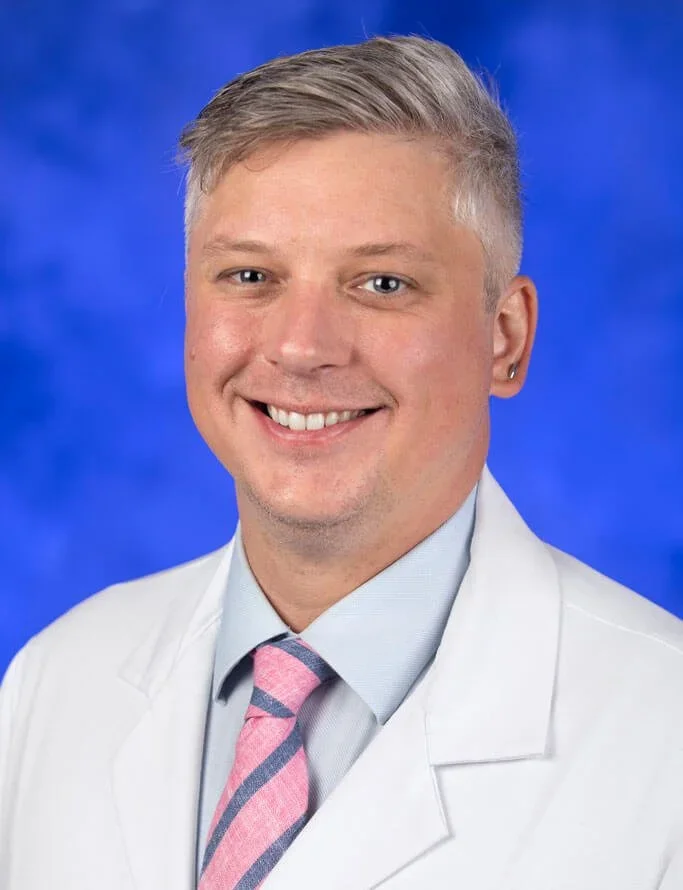 Portrait of a smiling young male doctor with short blond hair, wearing a white coat, light blue shirt, and a pink striped tie, with a blue background.
