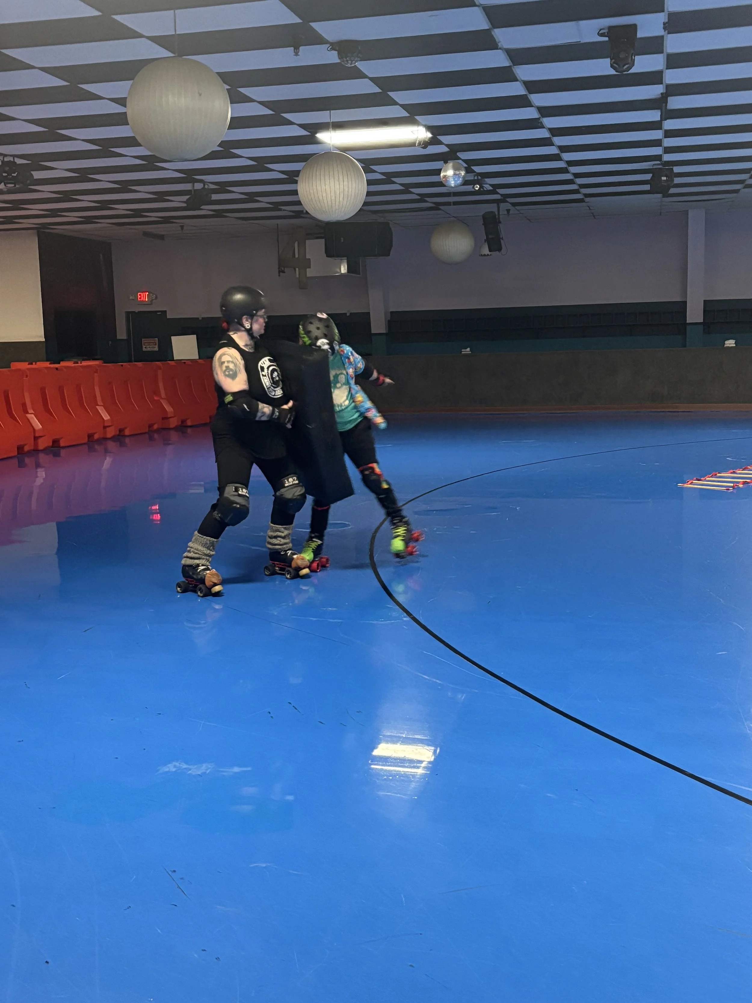 Two people with roller skates and helmets practicing roller derby in an indoor rink with a blue floor and red safety barriers.