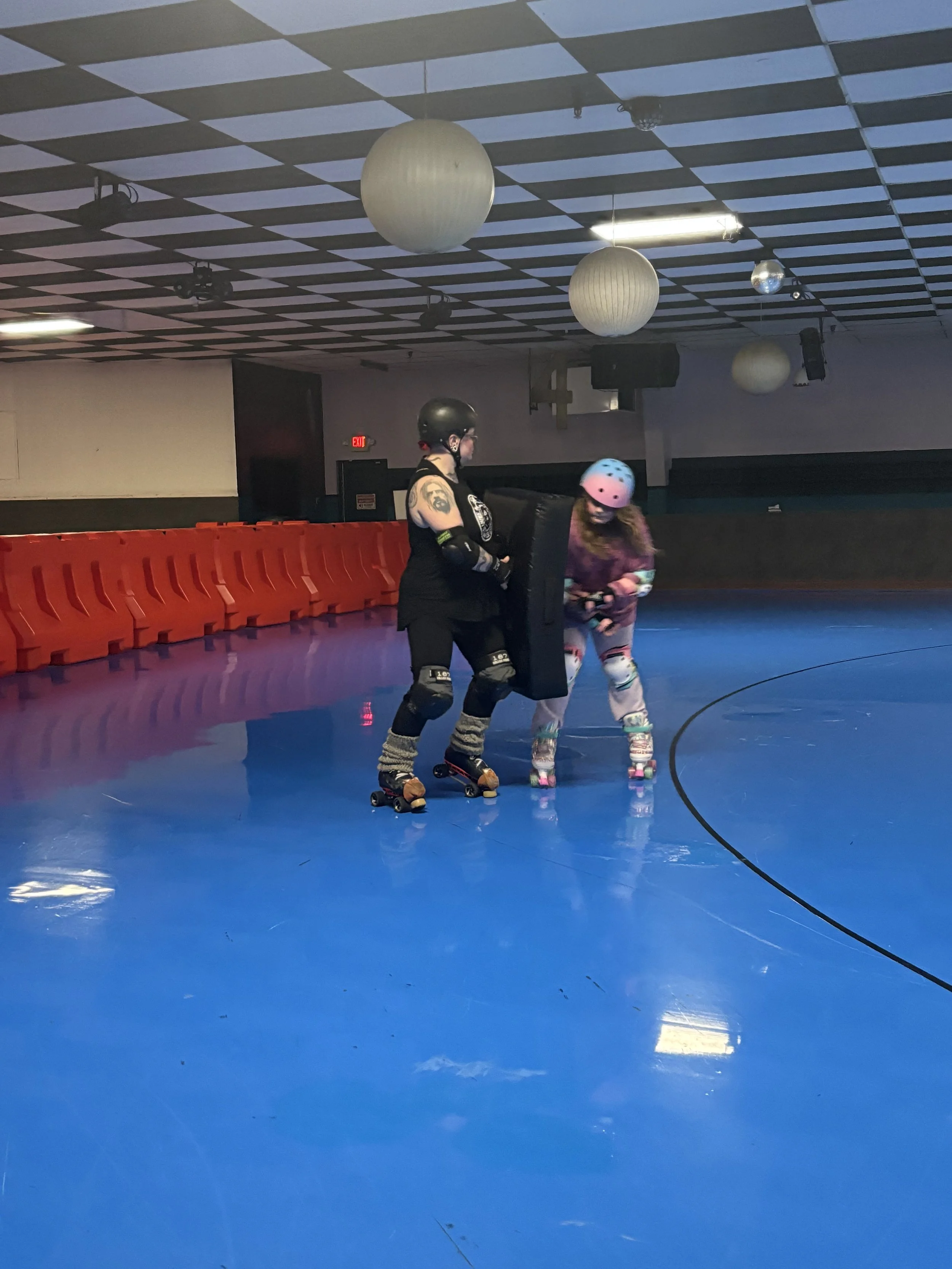Two women roller skating in an indoor rink, wearing protective helmets and pads, with one holding a large black padded shield, the other smiling while looking at her, a row of orange safety barriers, and a ceiling with paper lanterns and disco balls.