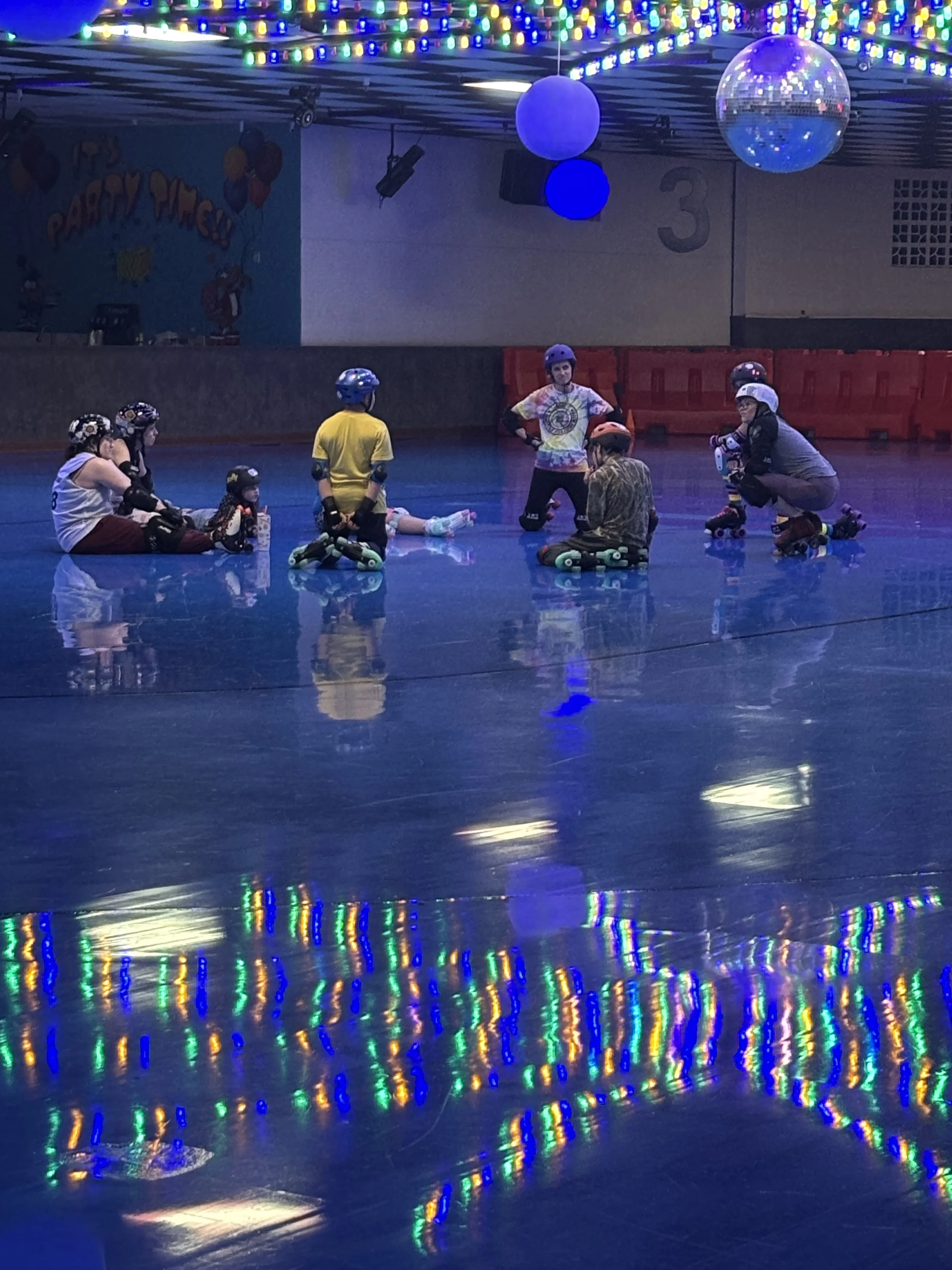 A group of children and adults roller skating in an indoor rink, with colorful lights and a disco ball overhead.