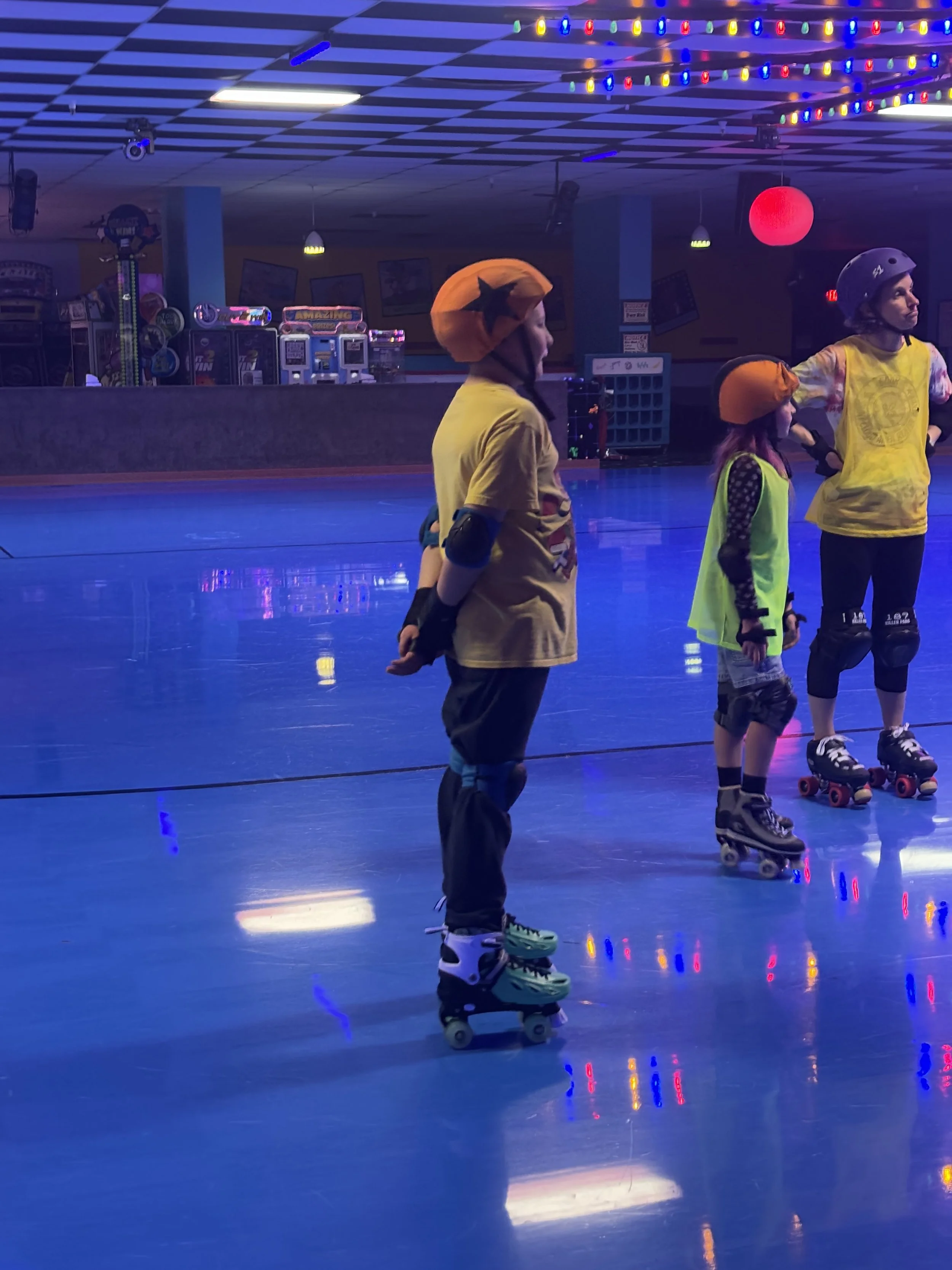 Three children in yellow shirts and helmets roller skating in an indoor rink with blue lighting and colorful string lights overhead; one child wearing a star hat.
