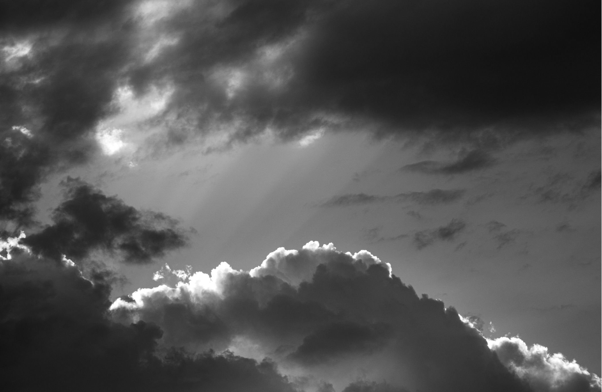 Black and white photograph of a cloudy sky with sunlight breaking through clouds in the background.