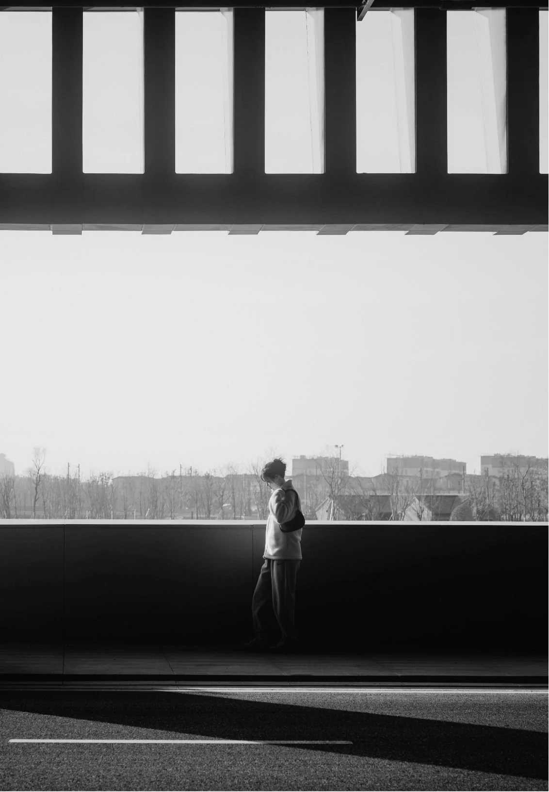 A person standing alone under an overpass in black and white, looking down while wearing a backpack, with a cityscape in the background.