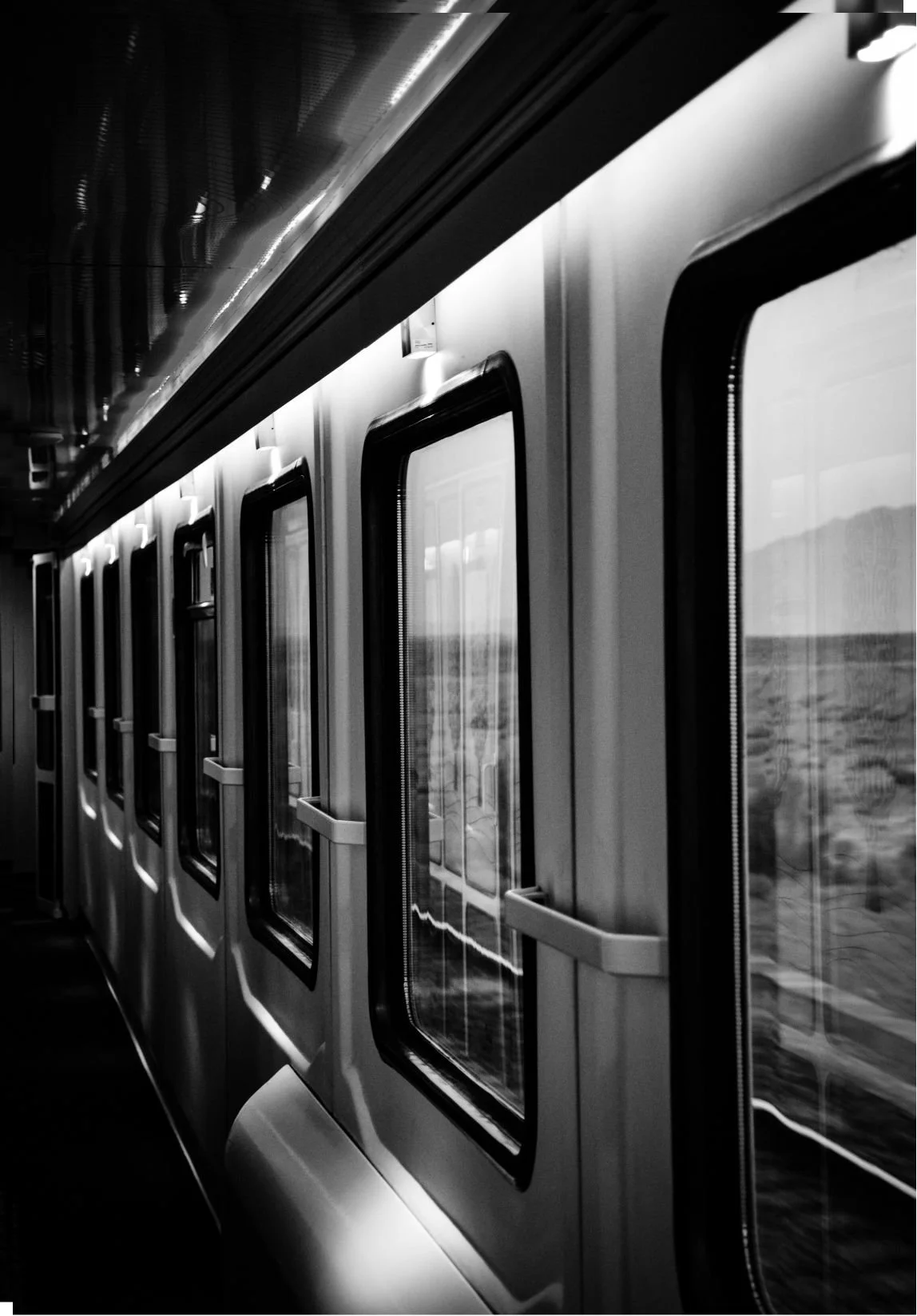 Black and white photo of a train interior showing multiple closed train windows along the wall, with reflections of the landscape outside visible through the windows.