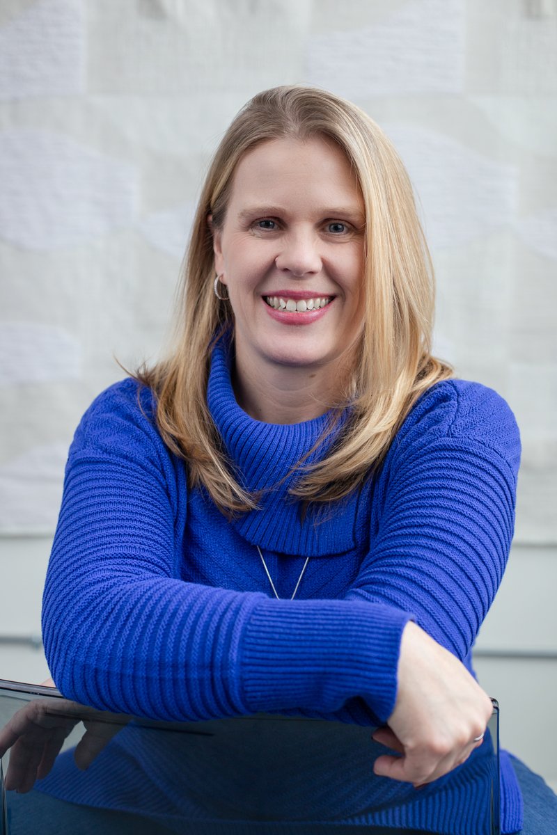 Ellie Feldmann, therapist for women, wearing a bright blue sweater, sitting at a desk with her arm resting on the surface, in an indoor setting.
