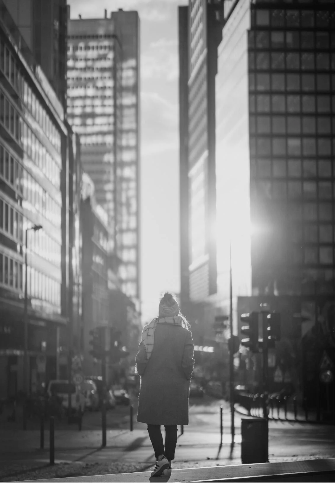 A woman walking in a city street surrounded by tall office buildings with glass windows, with sunlight shining through the buildings.