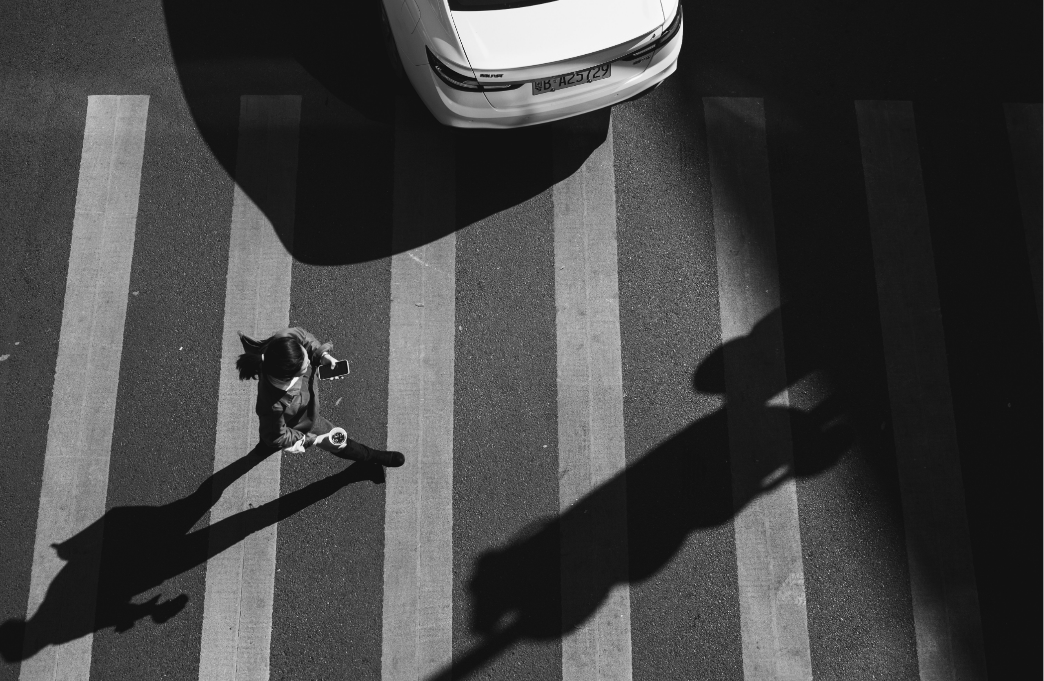 Black and white aerial view of a pedestrian crossing with a woman walking and holding a coffee cup and smartphone, casting a long shadow, and a parked car nearby.