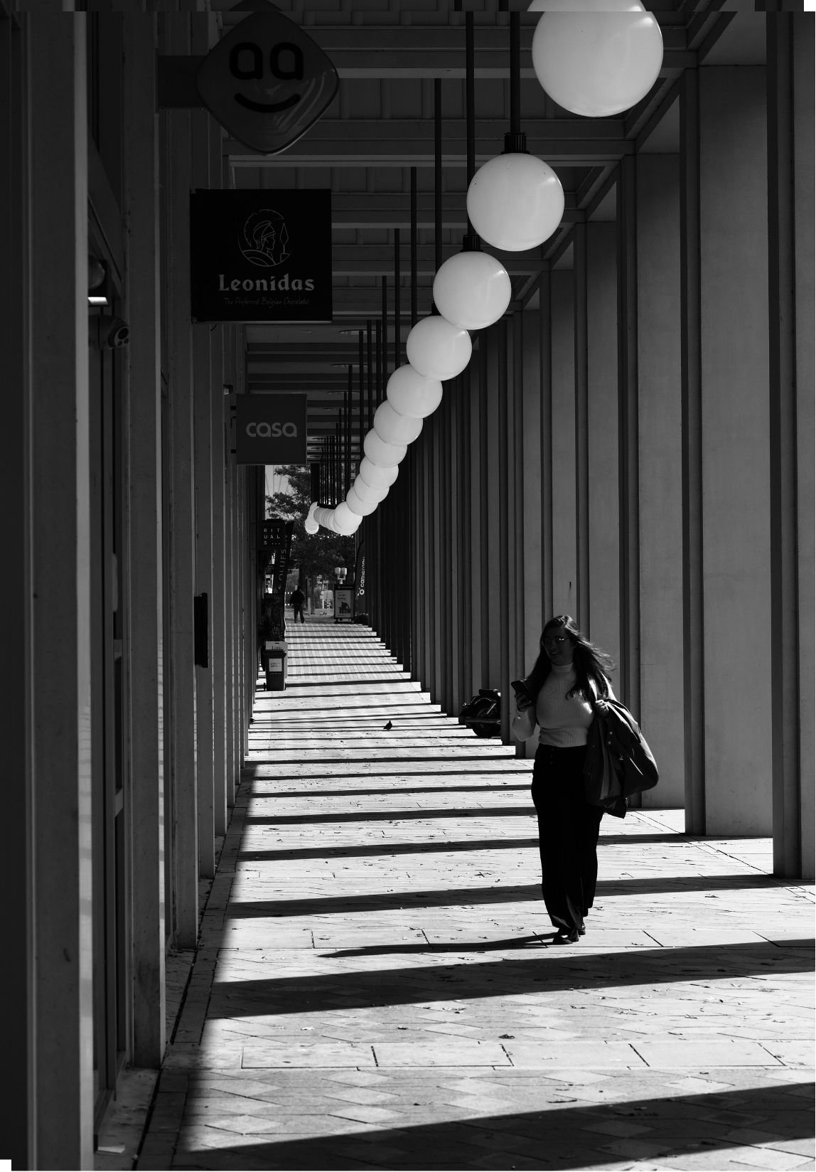 A woman walking on a covered outdoor walkway, illuminated by hanging spherical lights, with shadows cast on the ground.