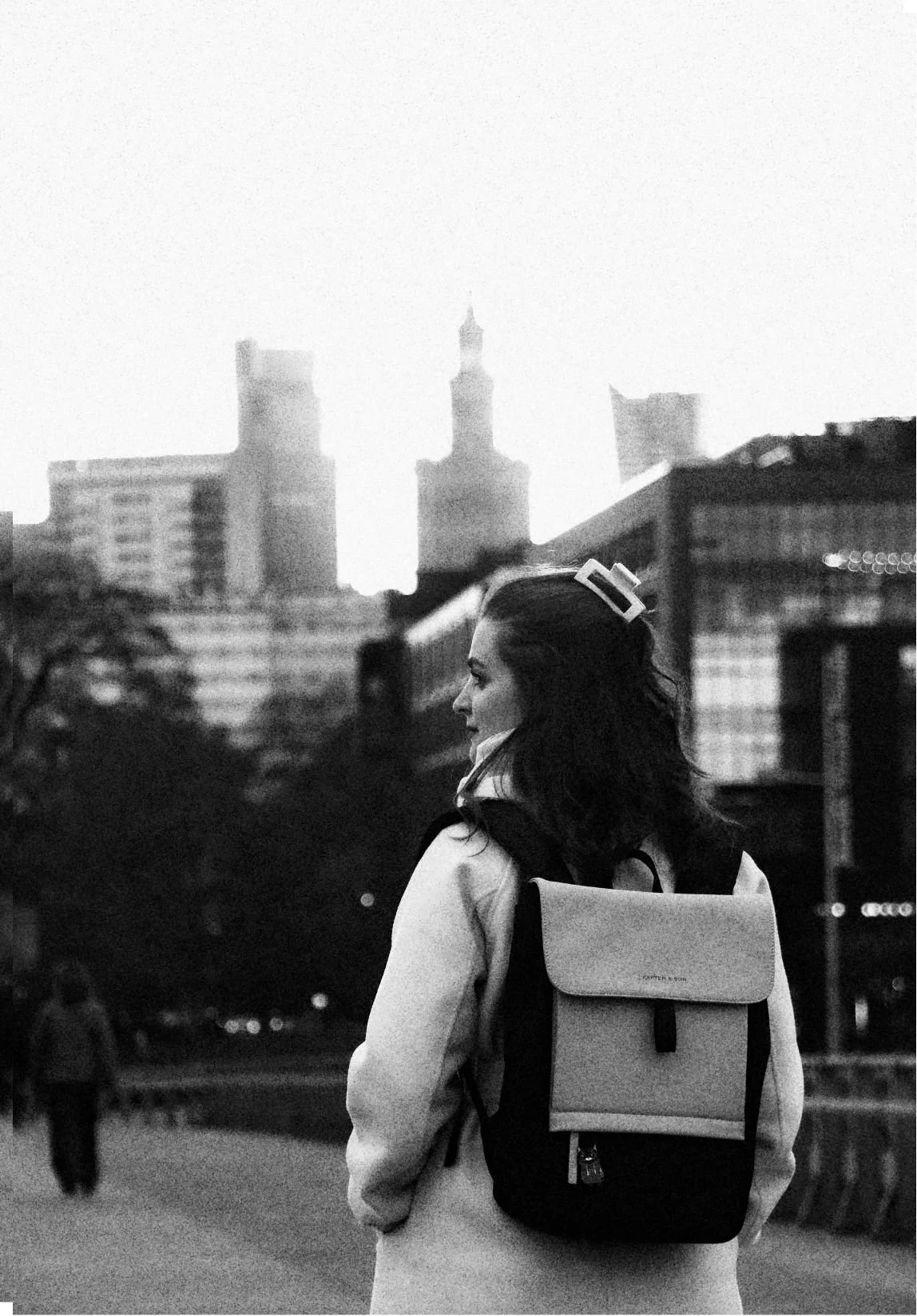 A woman with dark hair, wearing a coat and a backpack, standing outdoors in an urban setting with high-rise buildings in the background, black and white photo.