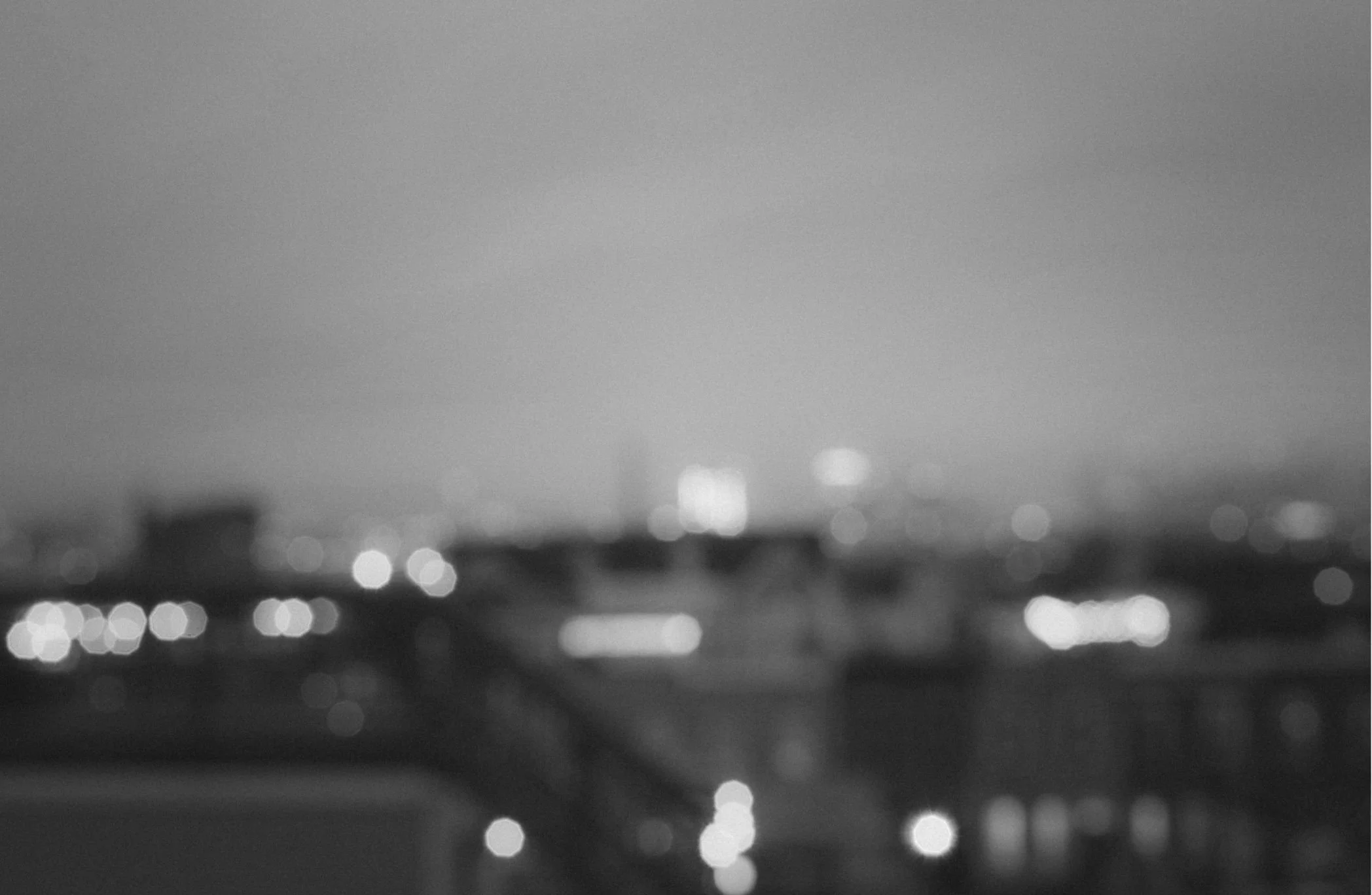 Black and white photo of a cityscape at dusk with blurred lights and buildings in the distance.