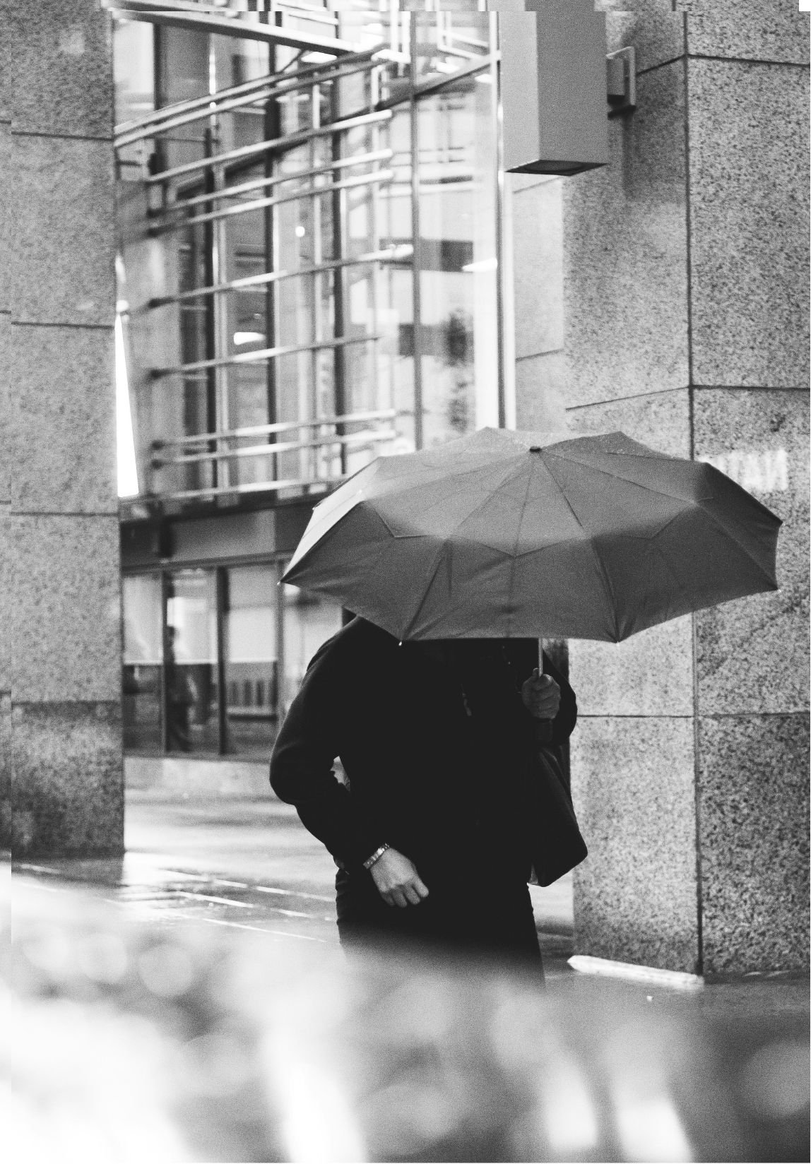 A person holding an umbrella walking on the street in front of a modern building with glass and stone walls on a rainy day.