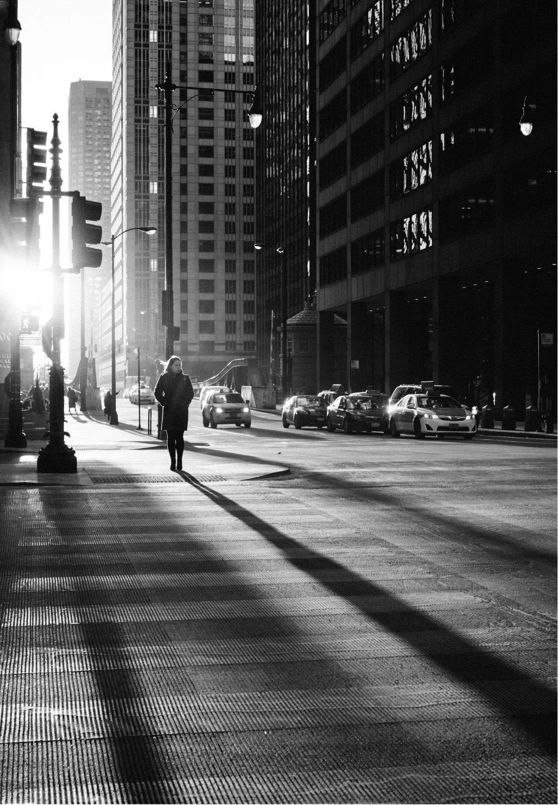Black and white photo of a city street during sunset with a person walking on the sidewalk and cars parked along the street.