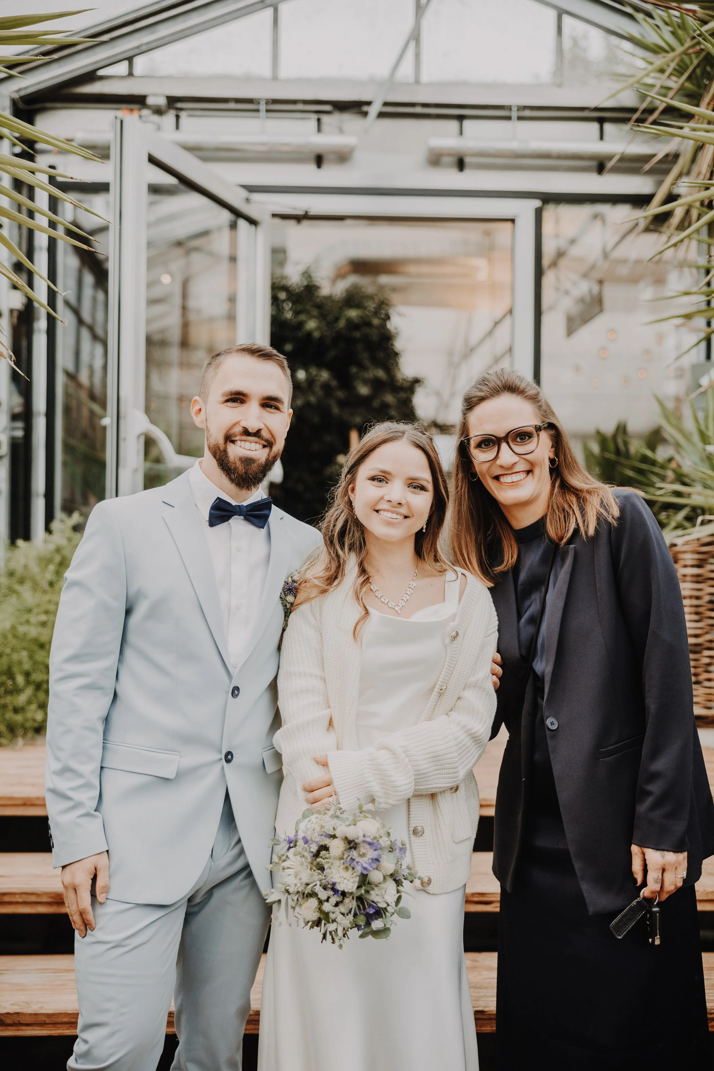 Drei Menschen, ein Brautpaar und eine Frau, stehen zusammen in einem Gewächshaus. Der Mann trägt einen weißen Anzug mit dunklem Fliege, die Frau eine weiße Brautkleid mit Schal, hält einen Blumenstrauß, die andere Frau trägt eine schwarze Jacke und Brille. Alle lächeln in die Kamera.